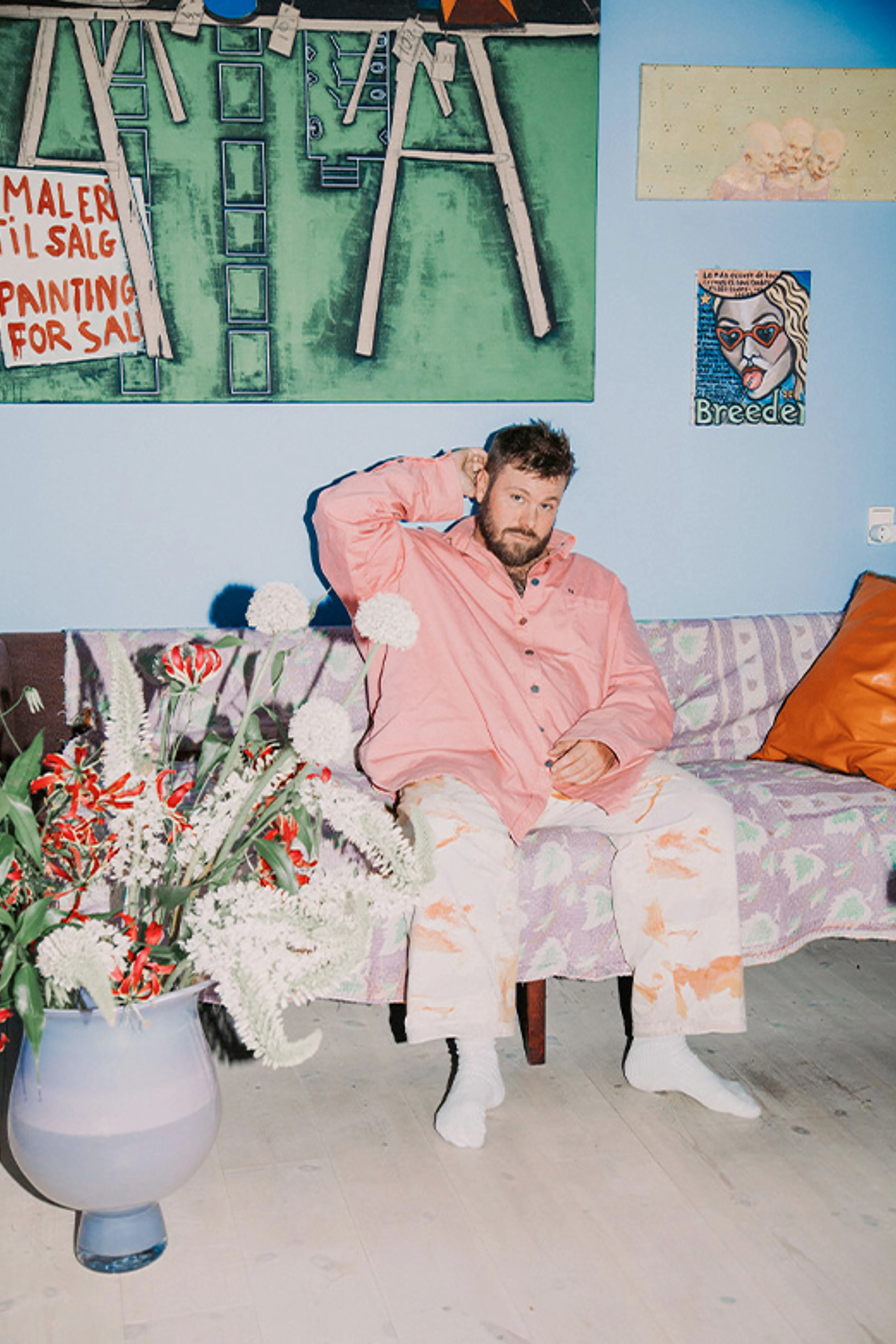 Mand i lyserød skjorte sidder på en mønstret sofa med en stor blomsterbuket og kunstværker omkring sig.