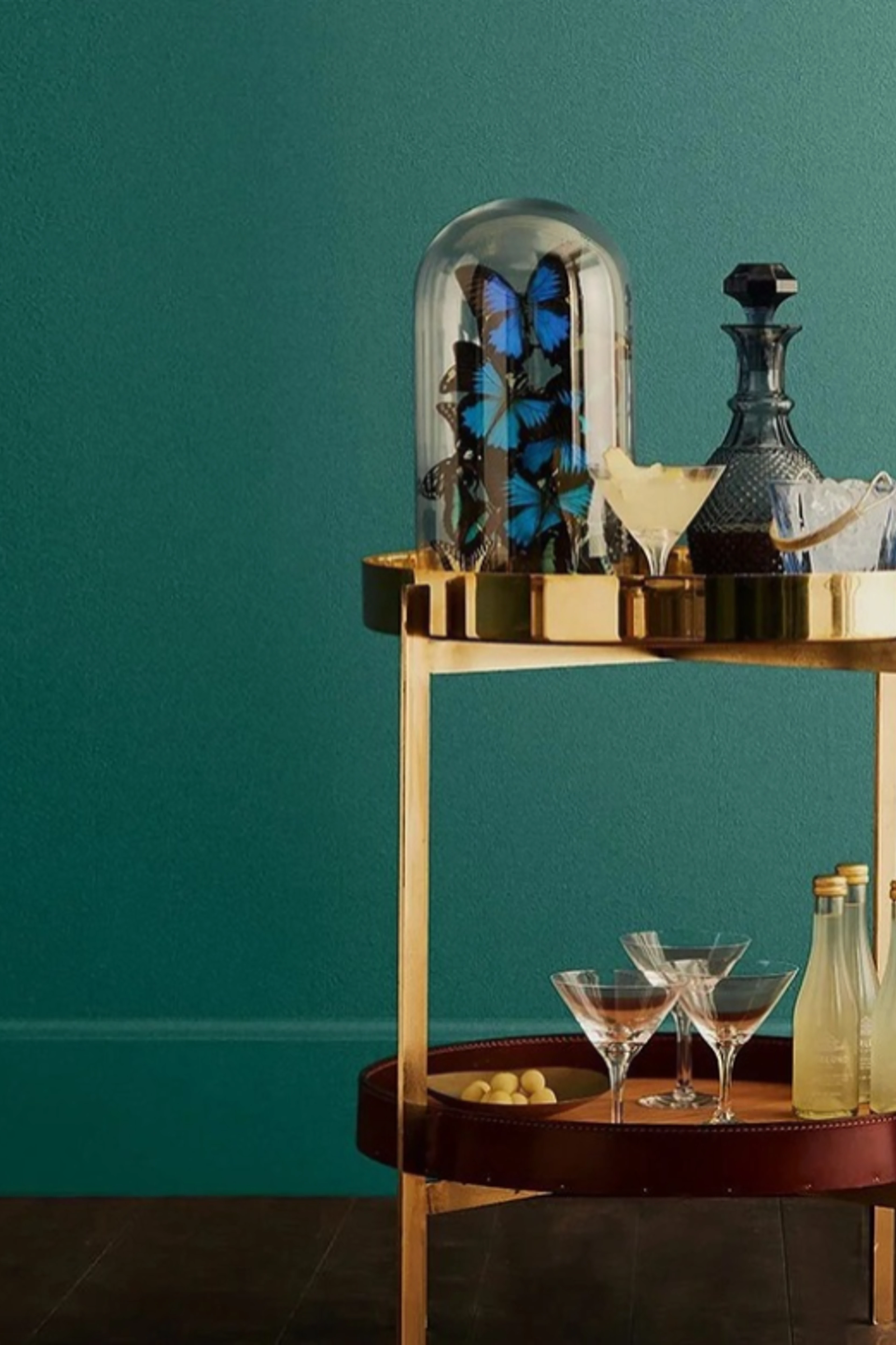 Petrolgrønn vegg med en gullfarget barvogn med cocktailglass, en karaffel og en glasskuppel med blå sommerfugler.
