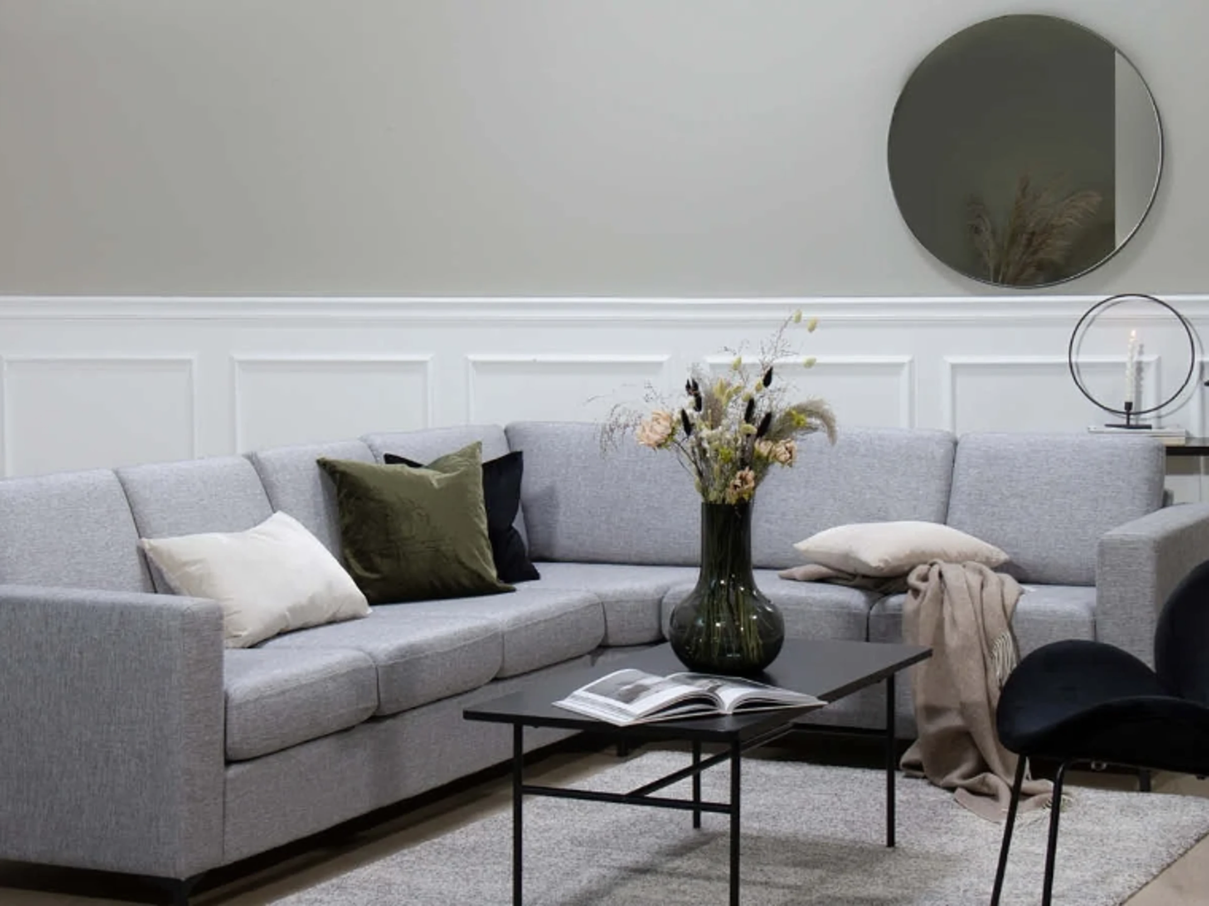 Stue med grå vegger og hvite veggpaneler, en lysegrå hjørnesofa med grønne og beige puter, et svart sofabord med en oppslått bok og en vase med tørkede blomster, et rundt speil og en svart stol.