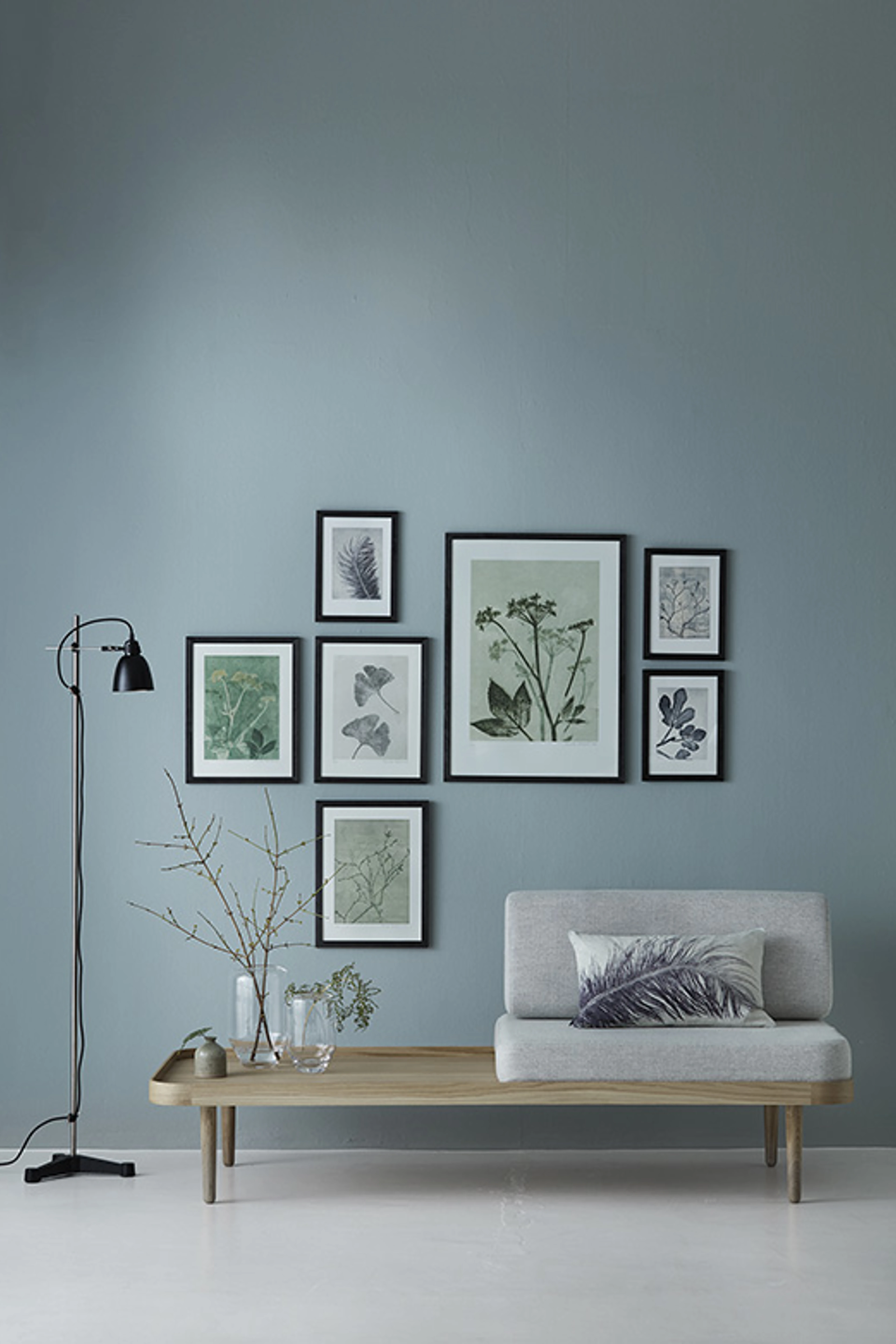 Stue med blågrå væg, lav bænk med puder, sort gulvlampe og en billedvæg med botaniske motiver i sorte rammer.
