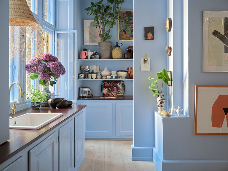 Lyst køkken i lyseblå nuancer med åbne hylder, planter og store vinduer.