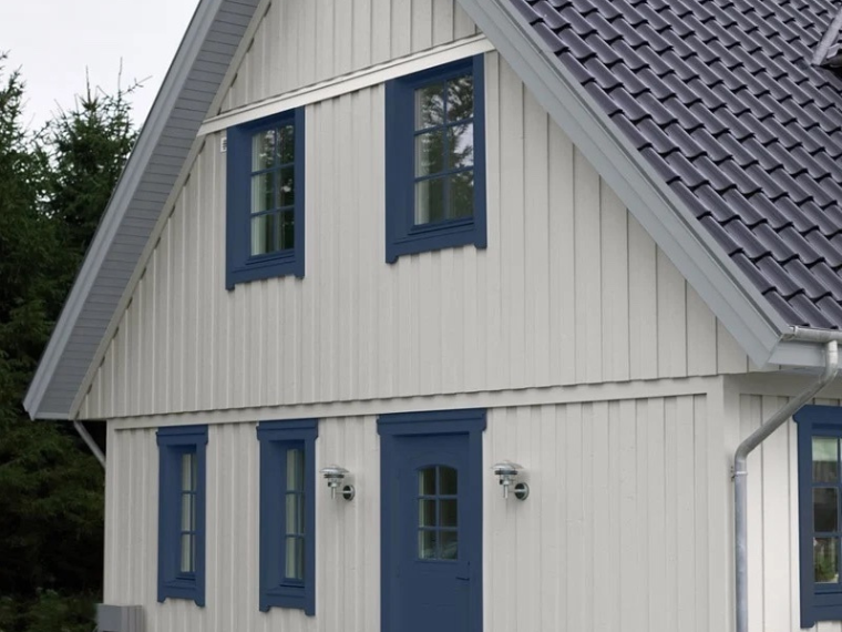 Vitt trähus med blå fönsterkarmar och dörr samt svart tegeltak, omgivet av barrträd.