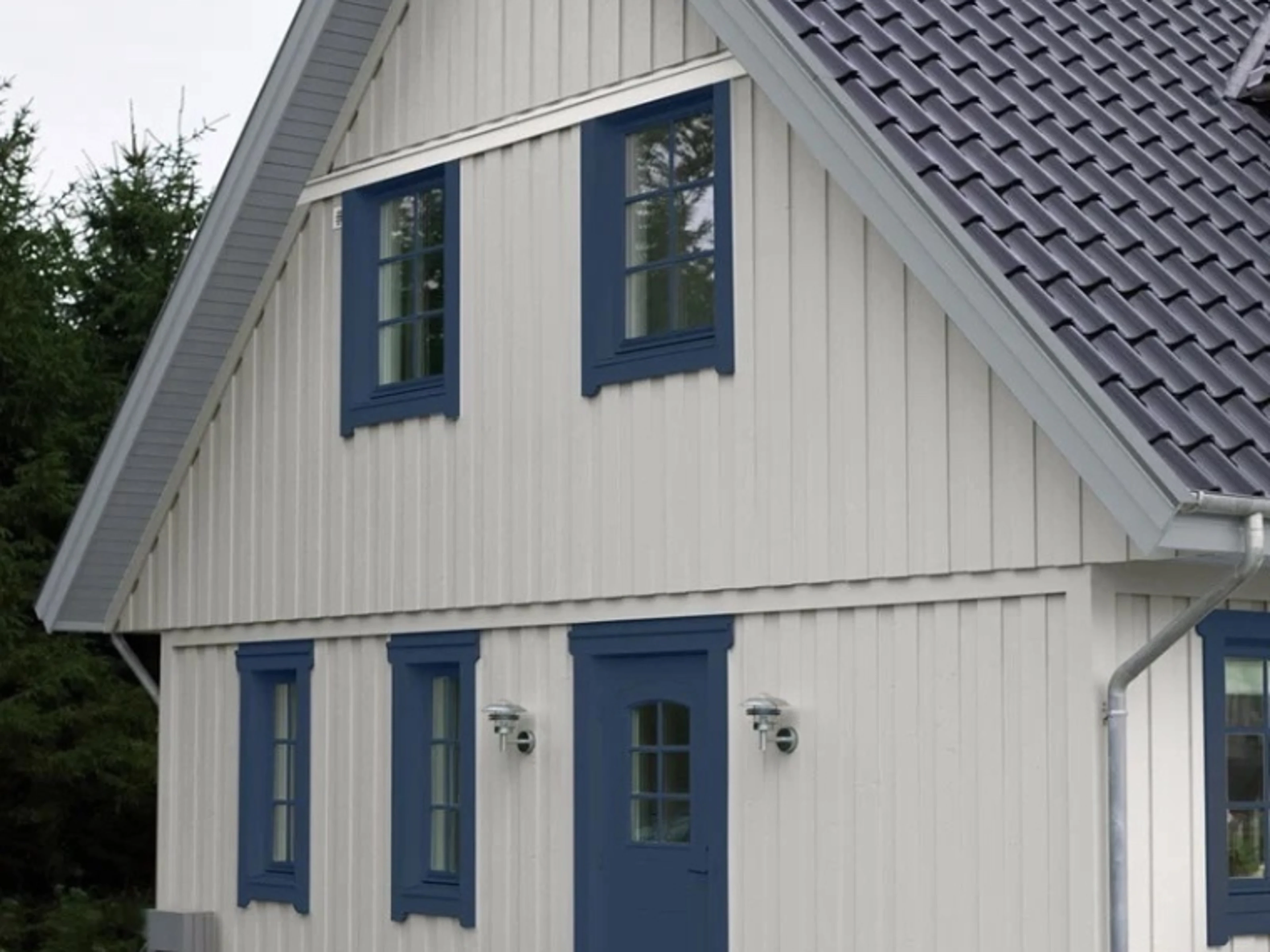Vitt trähus med blå fönsterkarmar och dörr samt svart tegeltak, omgivet av barrträd.