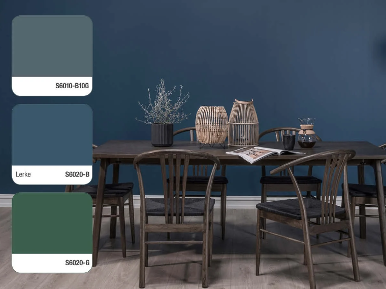 Dining area with dark blue walls, a dark wooden table with wooden chairs, rattan lanterns, a plant in a dark planter and an open magazine, with three colour swatches on the left: S6010-B10G, Lerke S6020-B and S6020-G.