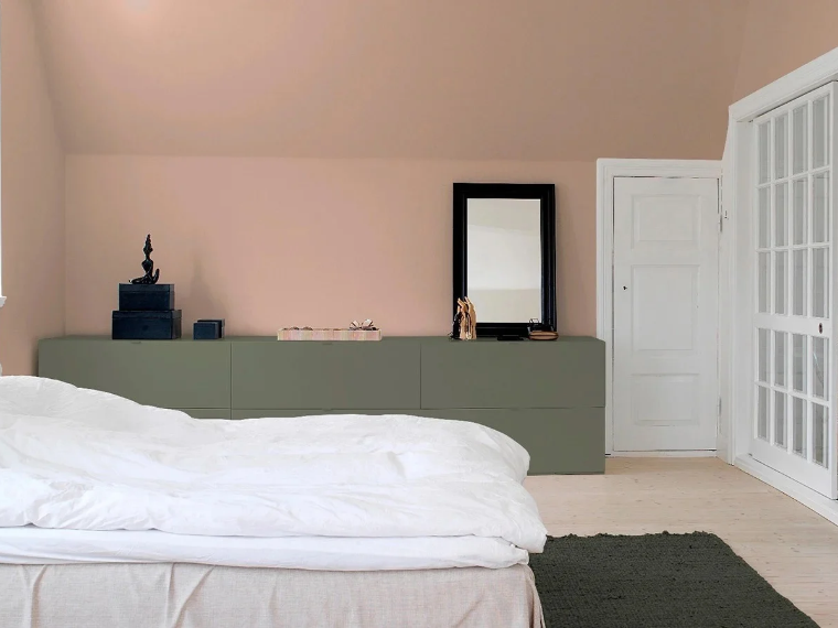 Soverom med støvete rosa vegger og skråtak, en seng med hvitt sengetøy, en lang olivengrønn kommode med et speil samt en hvit fyllingsdør og en glasskyvedør.