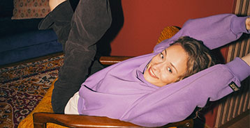 Kvinde i lilla sweatshirt læner sig afslappet tilbage i en sennepsgul lænestol med armene bag hovedet.