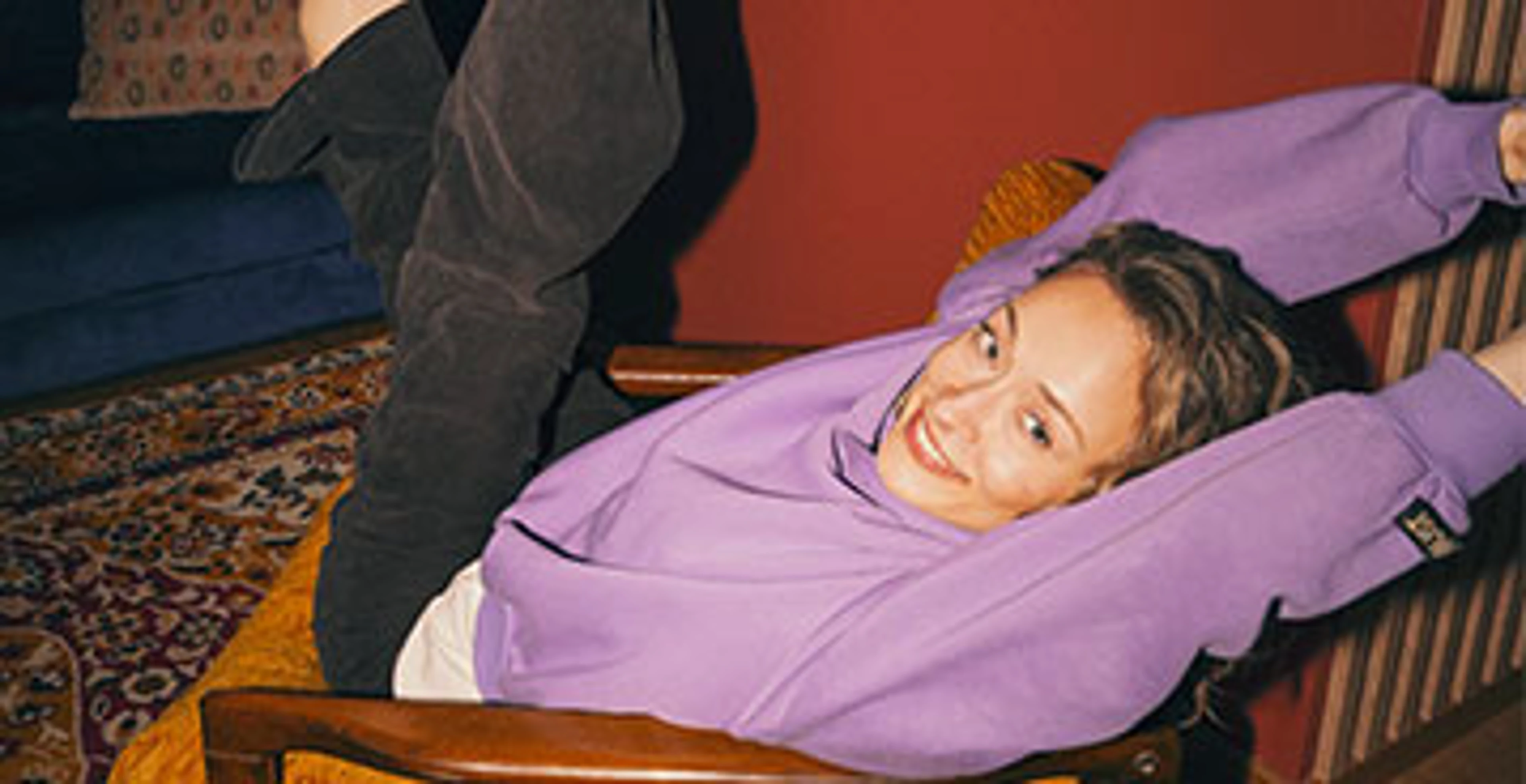 Kvinde i lilla sweatshirt læner sig afslappet tilbage i en sennepsgul lænestol med armene bag hovedet.