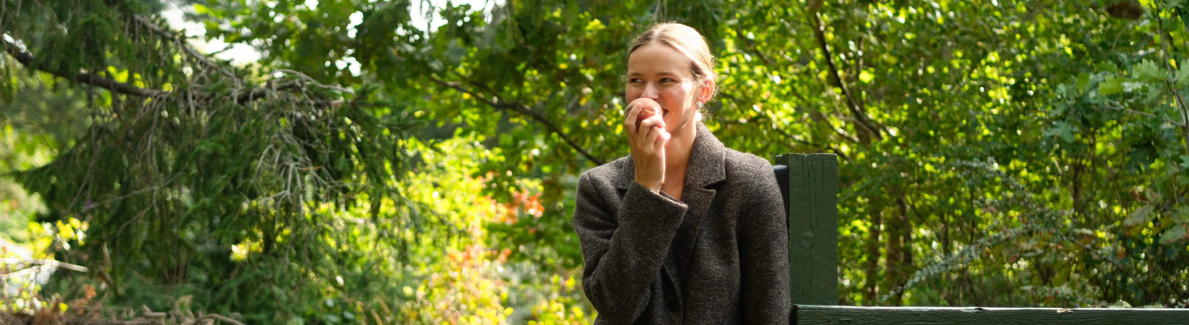 Woman eats an apple in the garden by a dark green fence.