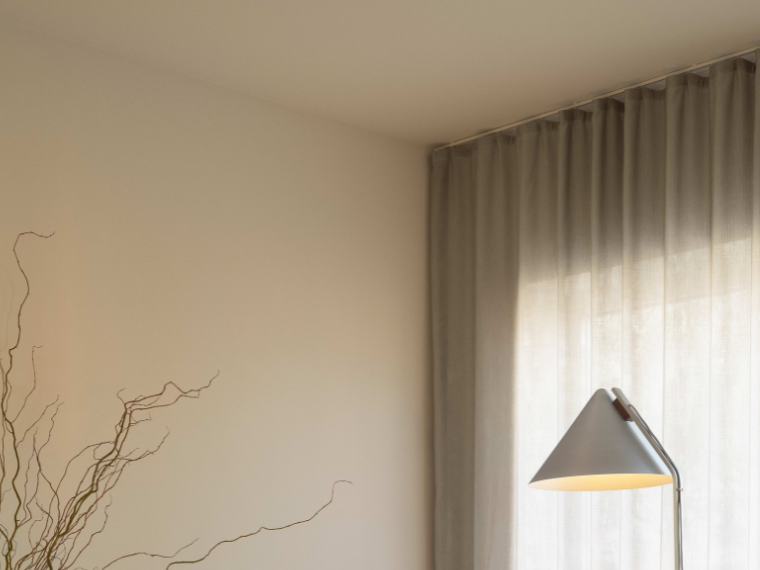 Lyst hjørne af en stue med gulvlampe, transparente gardiner og tørrede grene i forgrunden.