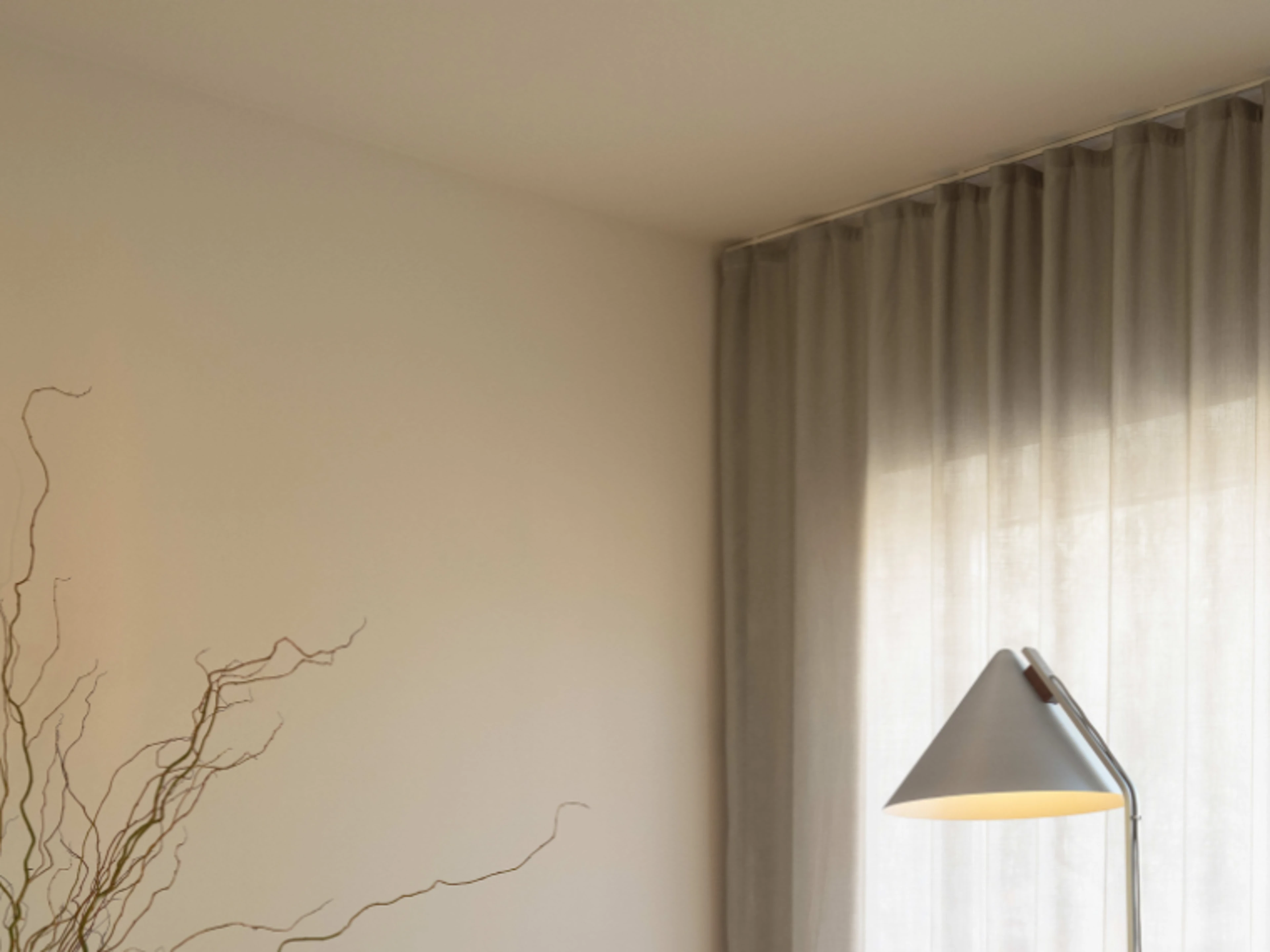 Lyst hjørne af en stue med gulvlampe, transparente gardiner og tørrede grene i forgrunden.