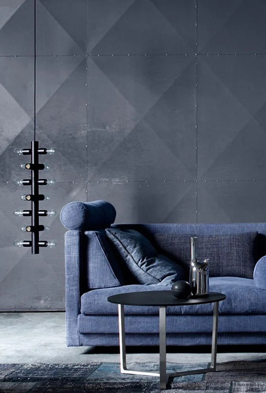 Stue med grå vægge, en blå sofa, og et sofabord med sort glasplade og metalben. Der er et blålig tæppe under sofabordet, og der hænger en aflang sort lampe med flere lyspære til venstre for sofaen.