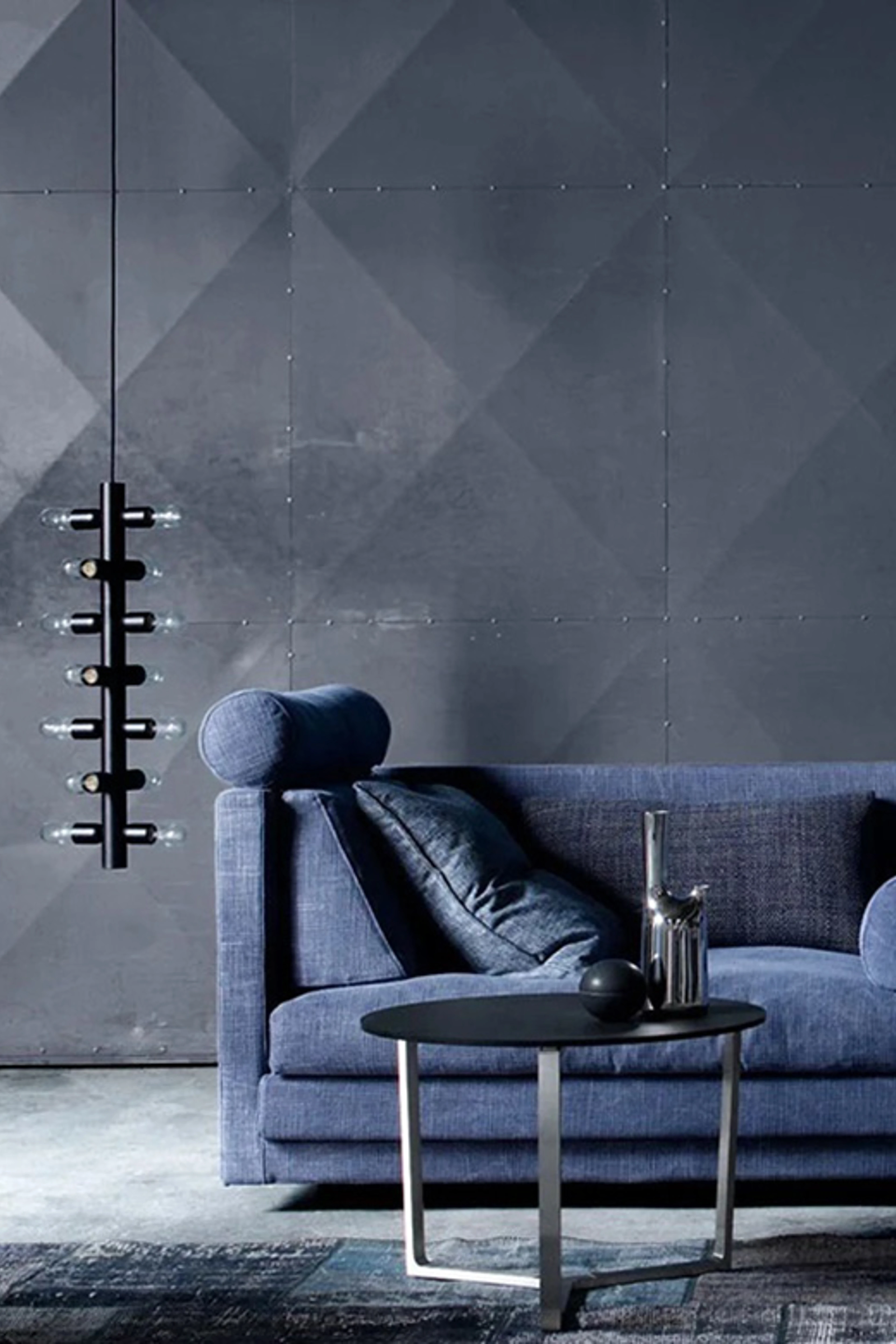 Stue med grå vægge, en blå sofa, og et sofabord med sort glasplade og metalben. Der er et blålig tæppe under sofabordet, og der hænger en aflang sort lampe med flere lyspære til venstre for sofaen.