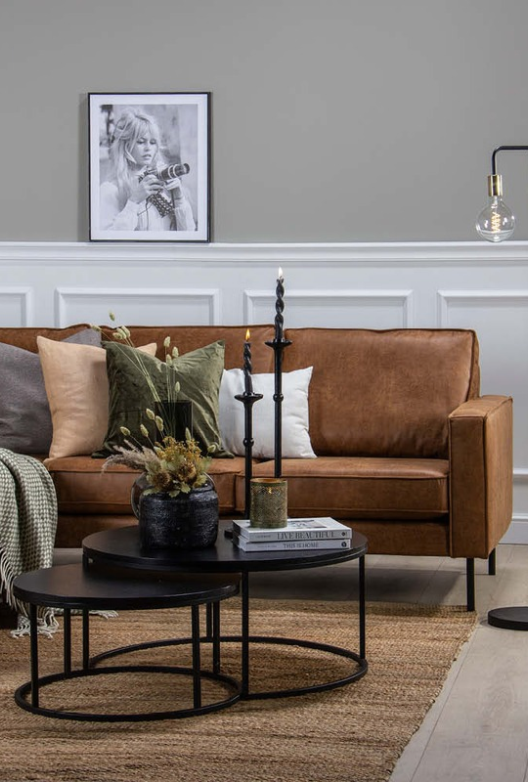 Stue med grå vegger og hvite veggpaneler, en cognacfarget skinnsofa med grønne og beige puter, runde svarte nesting-bord med lys og tørkede blomster, et juteteppe og et innrammet svart-hvitt fotografi.