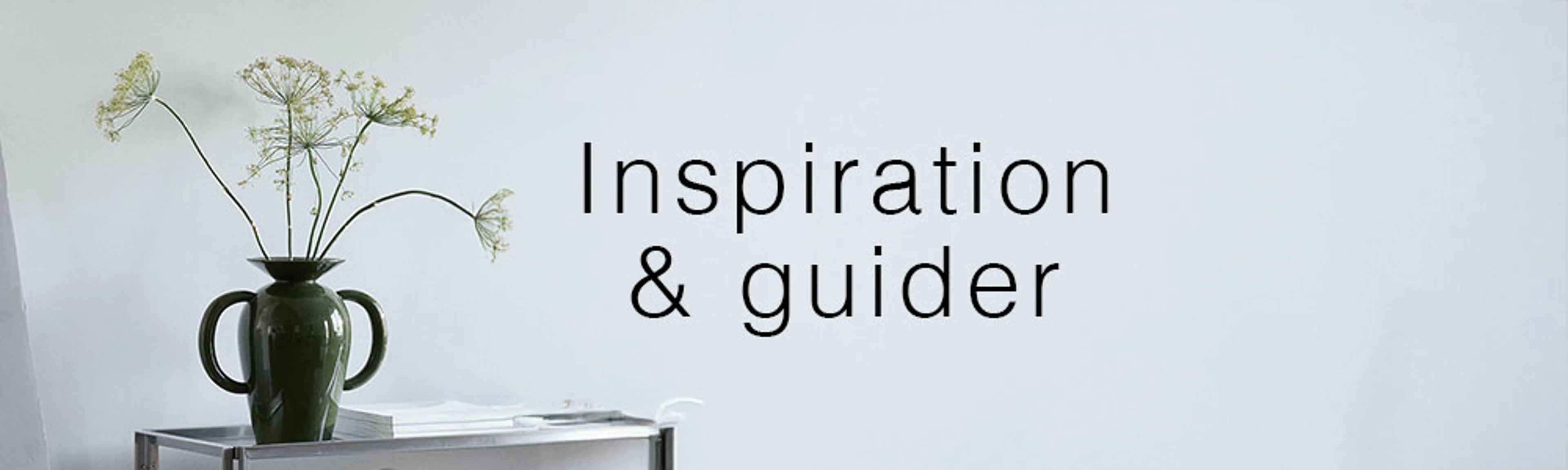 Vase med grønne blomster på et sidebord foran en lys væg med teksten "Inspiration & guider".