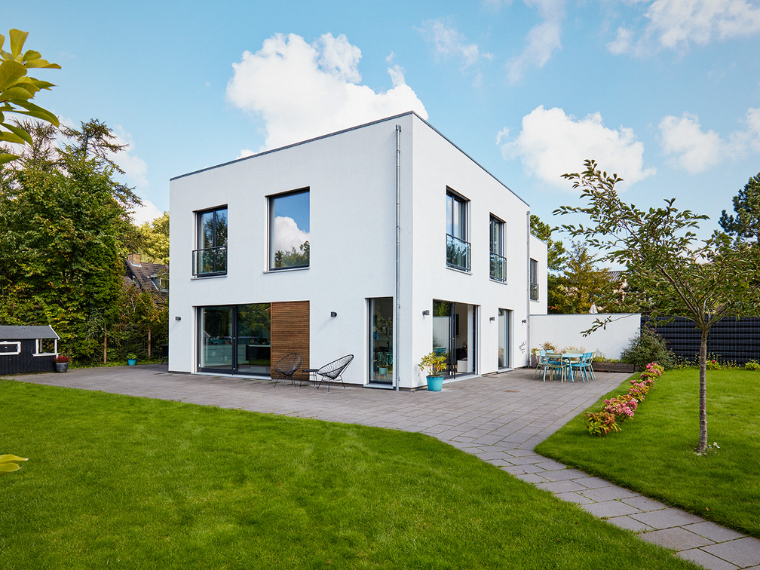 Modern white two-storey house with large windows, a paved terrace and a green garden.