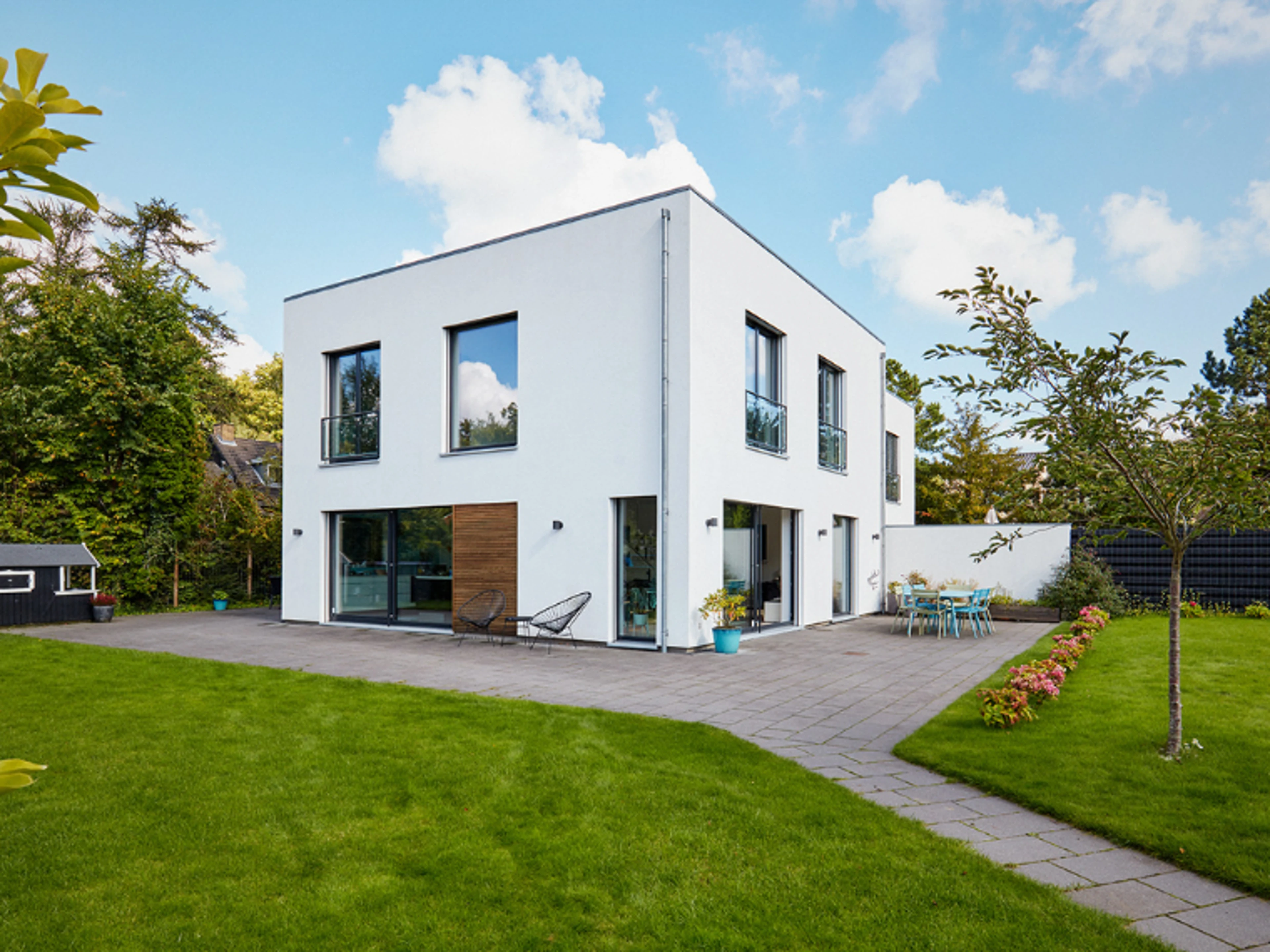 Modern white two-storey house with large windows, a paved terrace and a green garden.