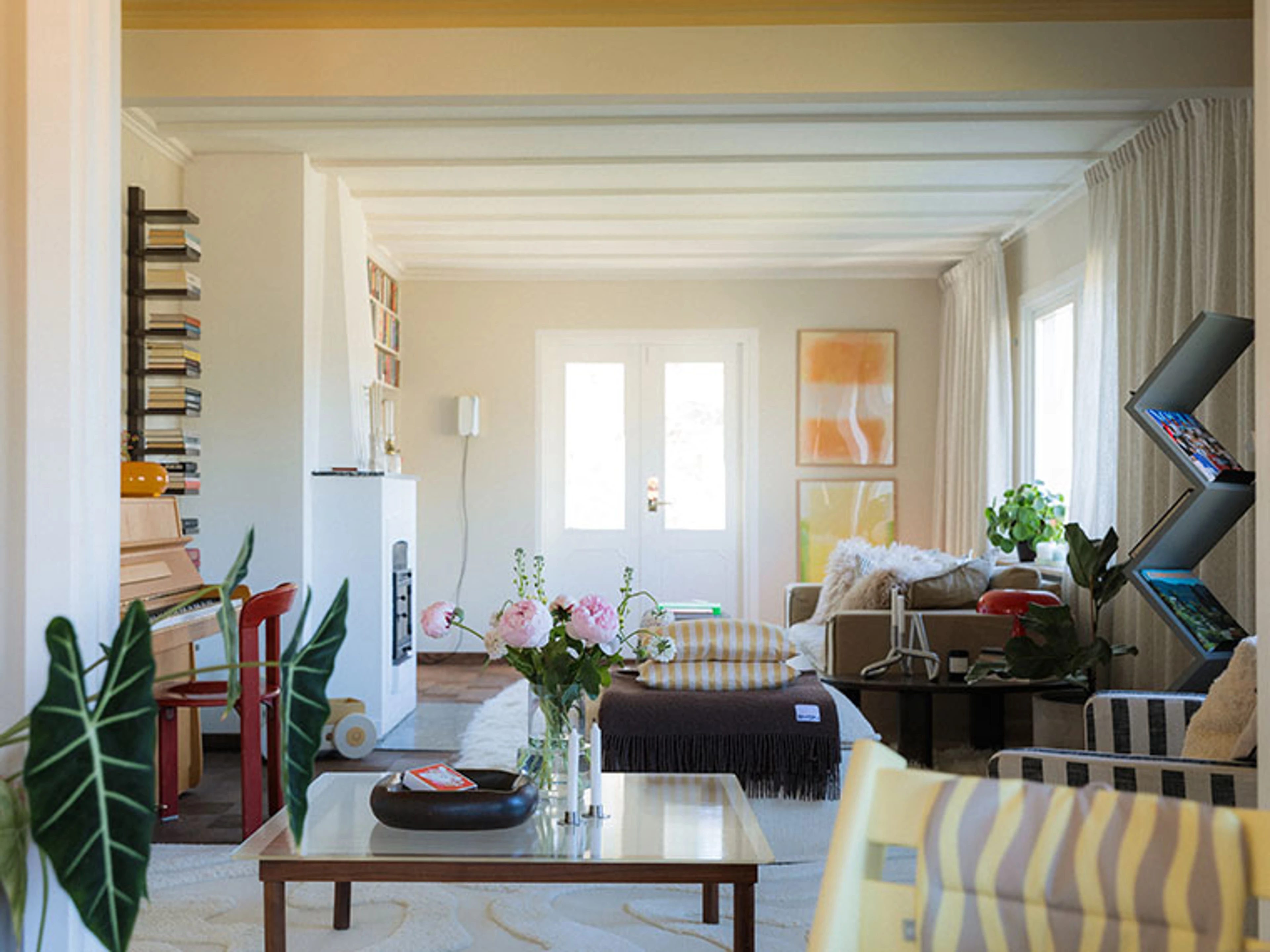 Living room with white ceiling beams, pink peonies and a piano in the background.