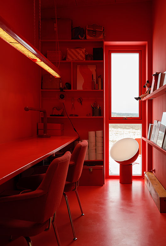 All-red home office with a long desk, red chairs, built-in shelves, and a narrow window.