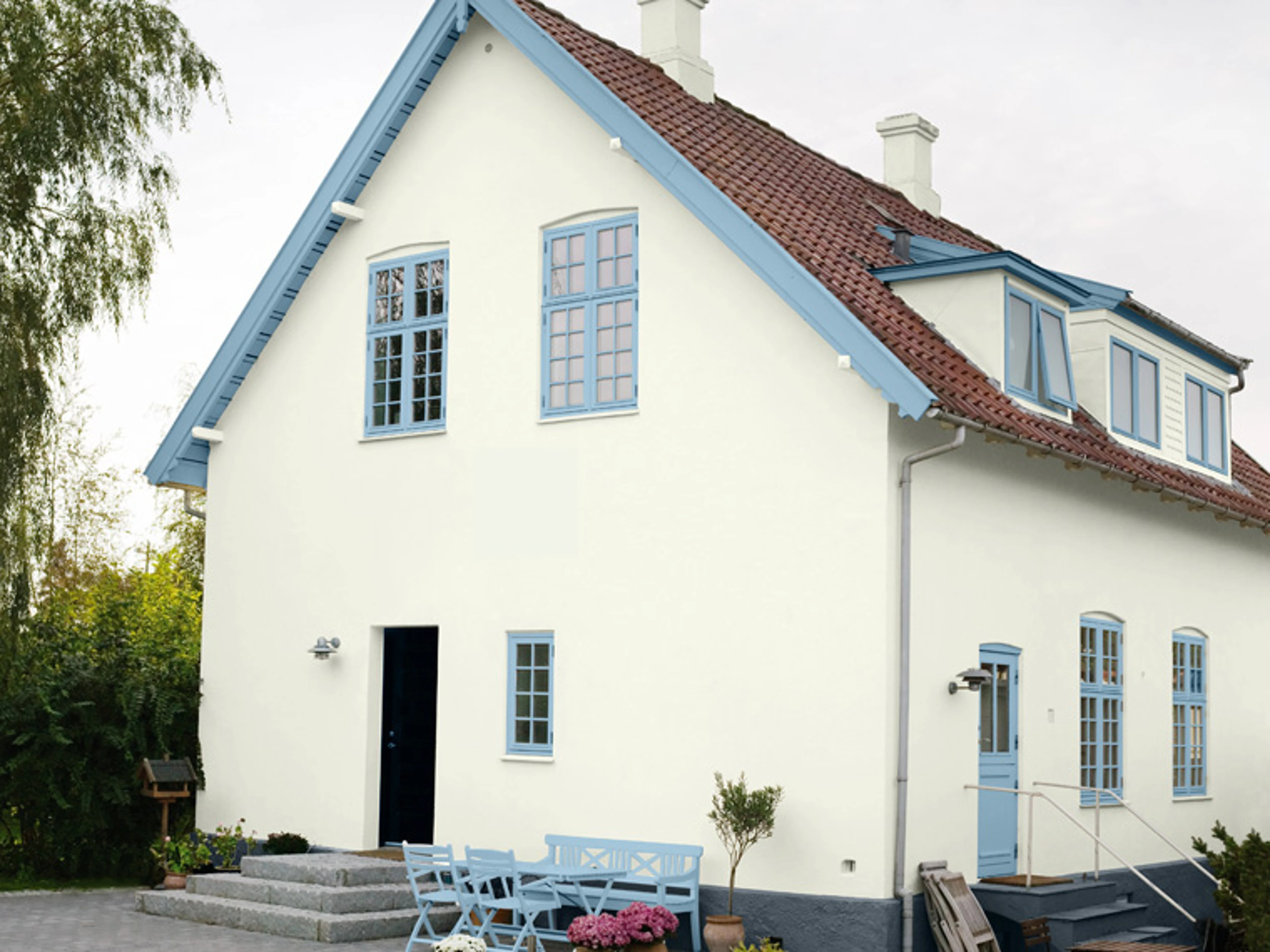 Vit husfasad med blå fönsterkarmar, rött tegeltak och matchande blå trädgårdsmöbler.