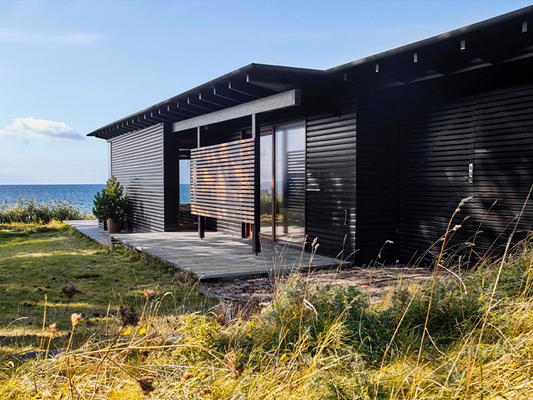 Moderne sommerhus med sortmalet træfacade og store glaspartier placeret tæt ved kysten. Huset er omgivet af marehalm og natur med havet i baggrunden.