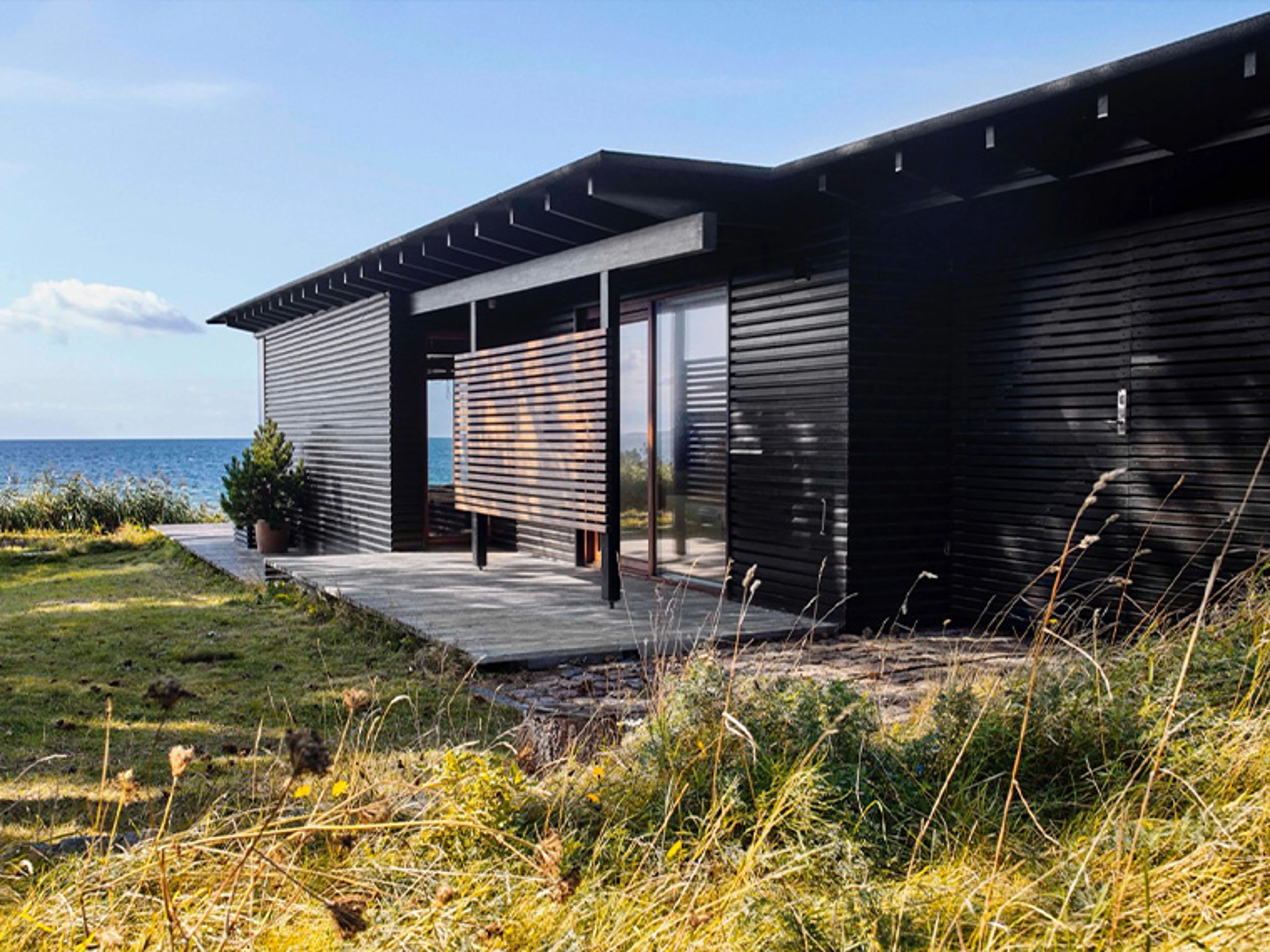 Moderne sommerhus med sortmalet træfacade og store glaspartier placeret tæt ved kysten. Huset er omgivet af marehalm og natur med havet i baggrunden.