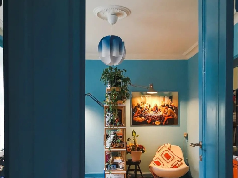 Rom med en klar blå vegg, sett gjennom en blå døråpning. Rommet inneholder en stol med mønstret pute, en bokhylle med planter og bøker, en gulvlampe samt et innrammet bilde på veggen. En blå pendellampe henger fra taket.