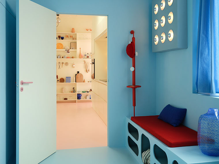 Blue entryway with a red bench, sculptural coat rack, and a view into a bright kitchen with a pink floor.