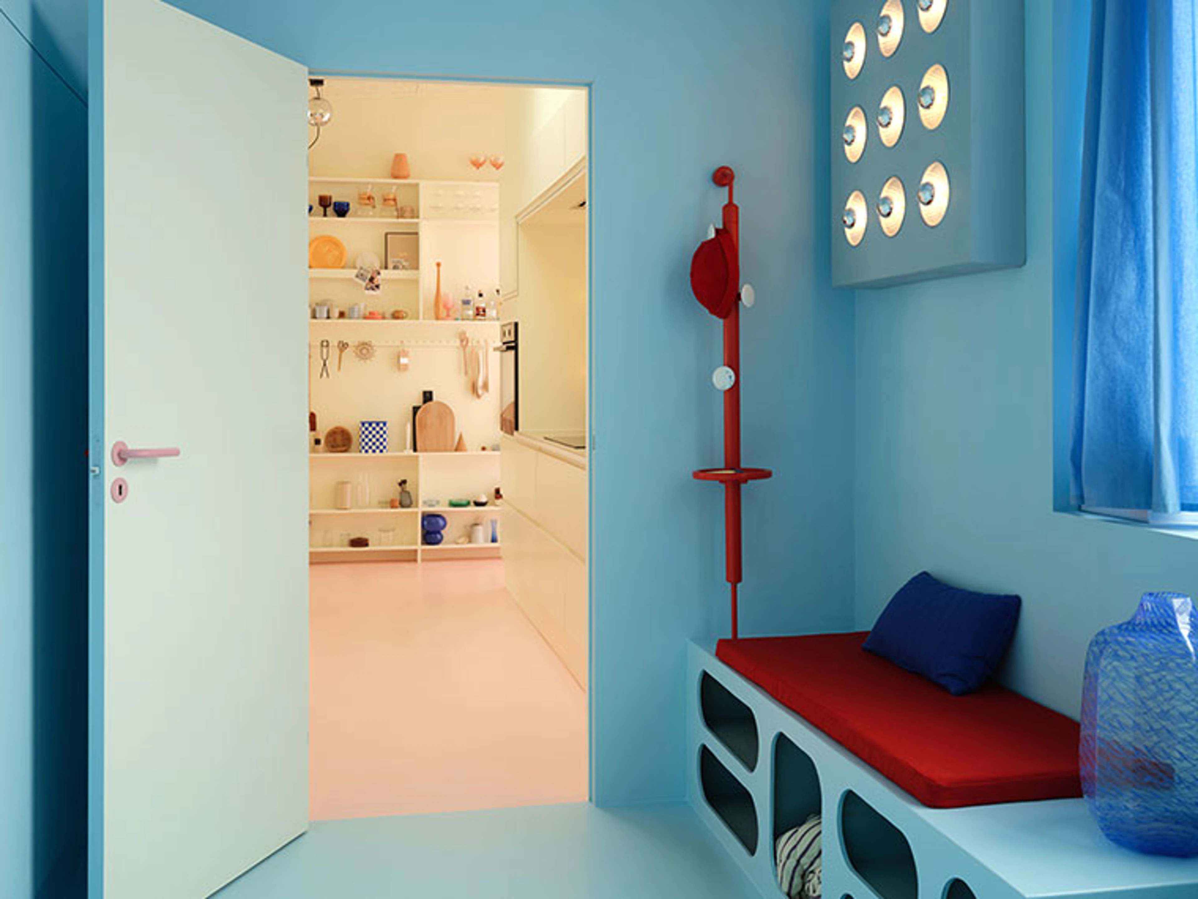 Blue entryway with a red bench, sculptural coat rack, and a view into a bright kitchen with a pink floor.