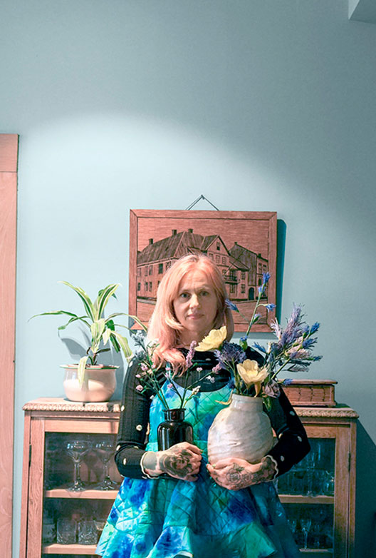 Kvinde med lyserødt hår står foran en lyseblå væg og holder to vaser med blomster foran et vintage vitrineskab.