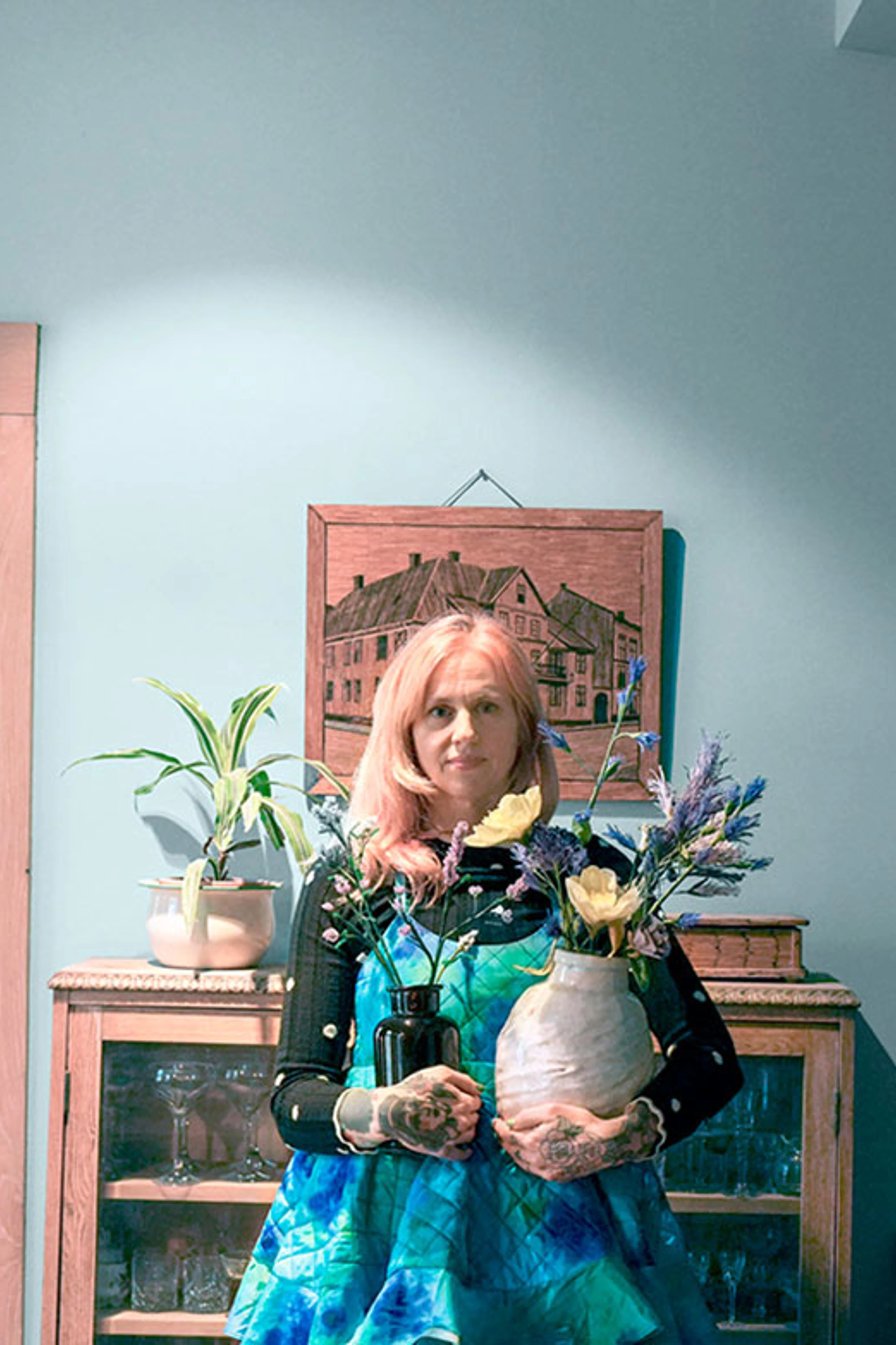 Kvinde med lyserødt hår står foran en lyseblå væg og holder to vaser med blomster foran et vintage vitrineskab.