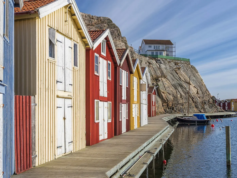 Färgglada sjöbodar i trä i rött, gult och blått längs en träbrygga vid vattnet, med en klippa i bakgrunden