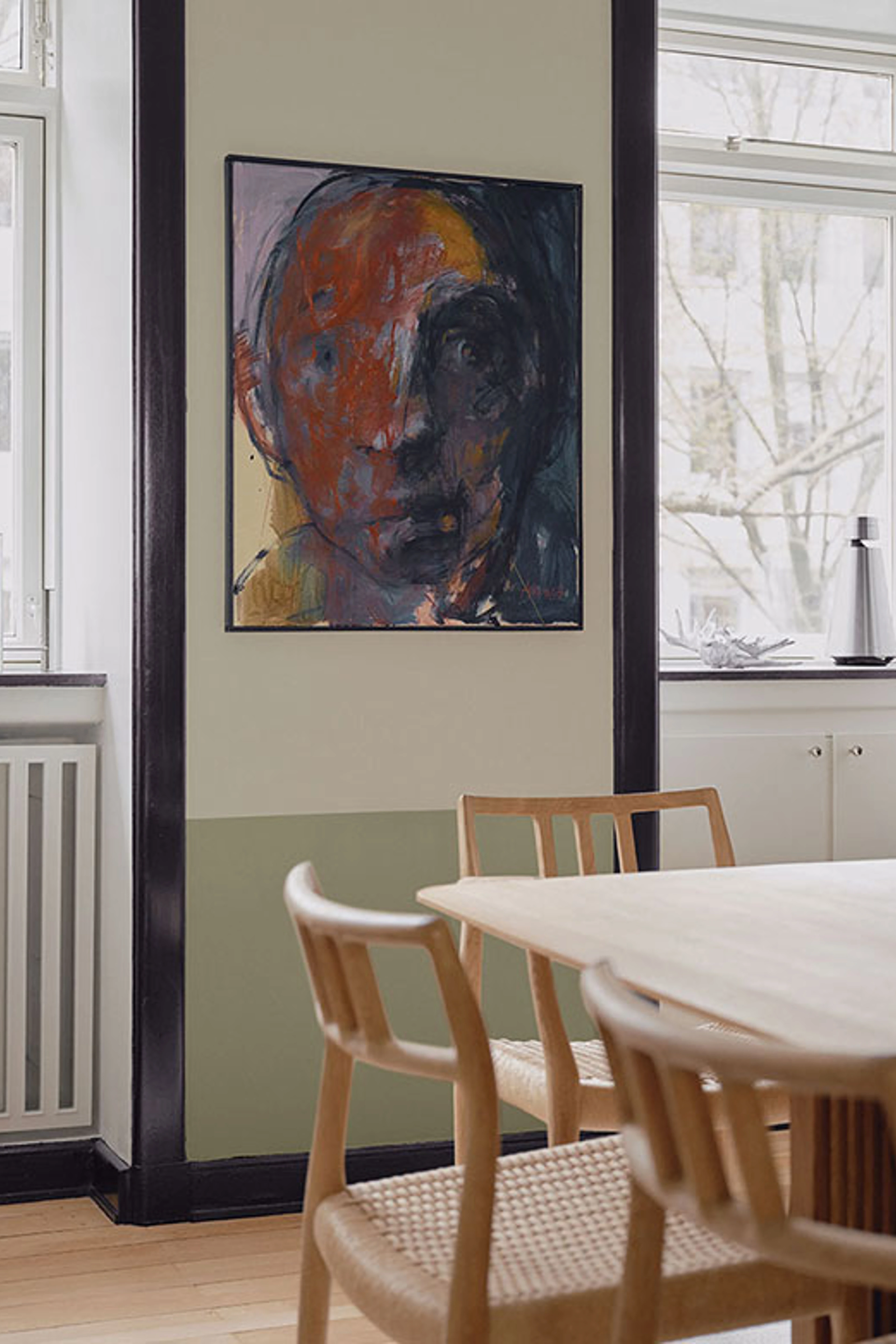 Væg i 2 grønlige nuancer i spisestuen med et abstrakt maleri af et ansigt i midten, omringet af sorte paneler.
