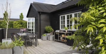 Hytte i tre med svart trebeskyttelse samt en treterrasse med mange planter.
