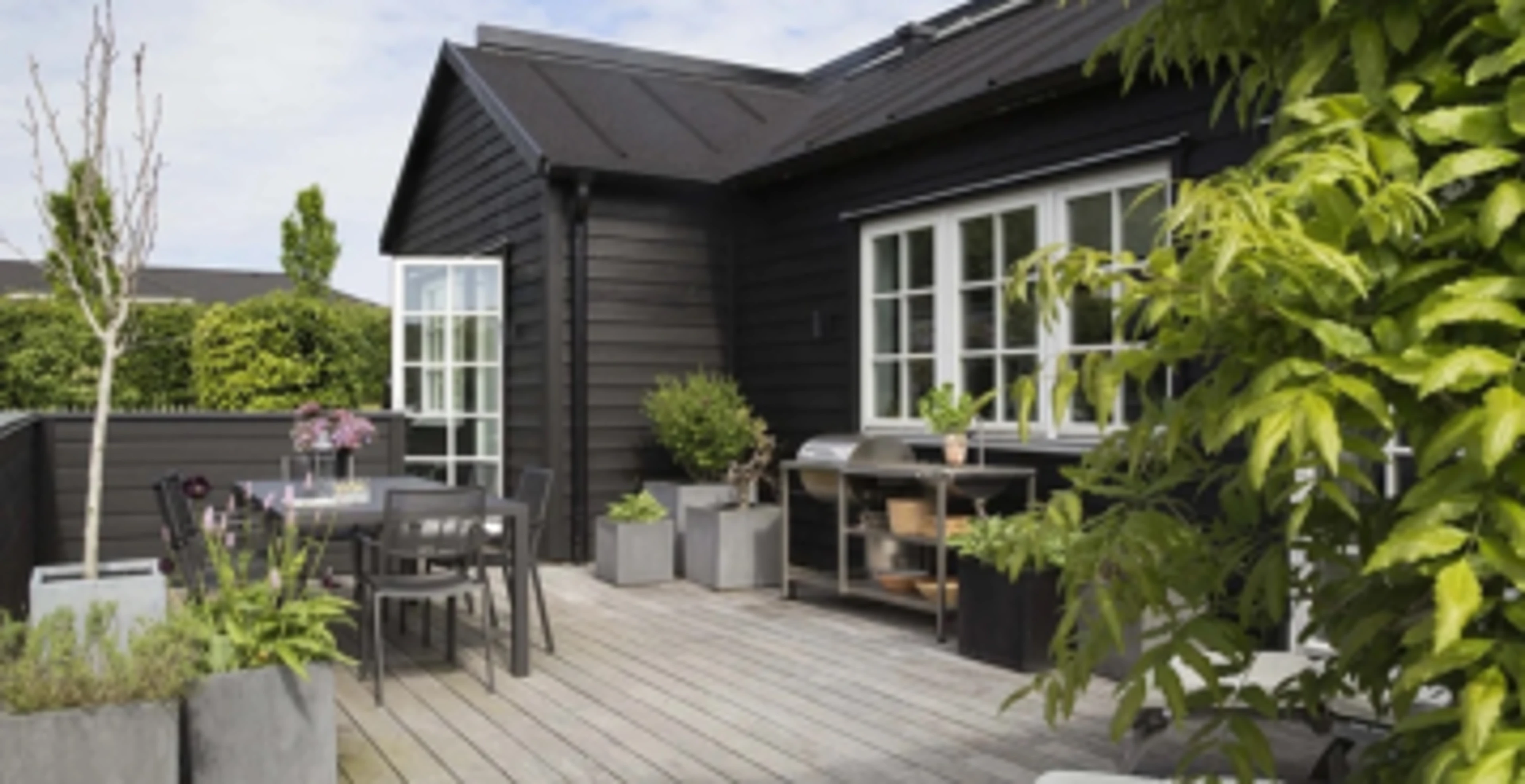 Hytte i tre med svart trebeskyttelse samt en treterrasse med mange planter.