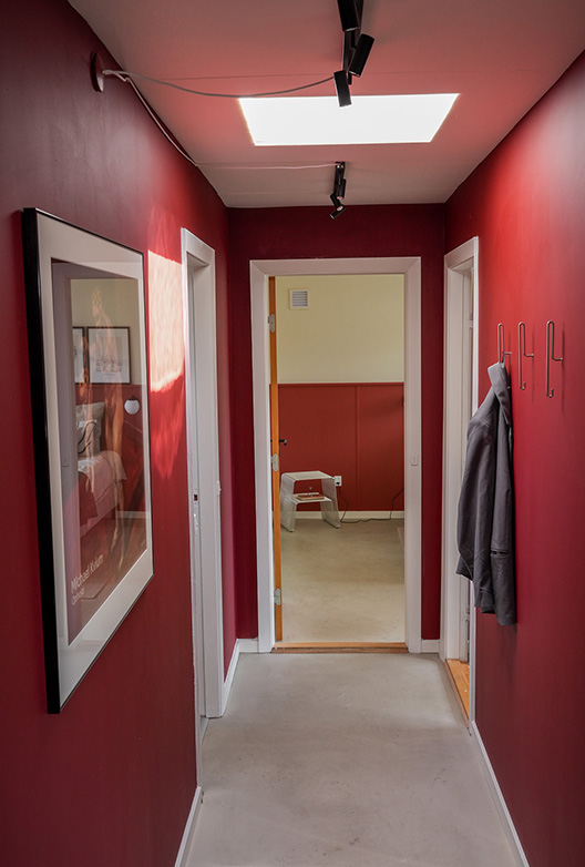 Smal gang med vægge malet i en dyb rød nuance. En indrammet plakat hænger på den ene væg, mens knager med ophængt overtøj og spotbelysning fremhæver rummets farverige udtryk.