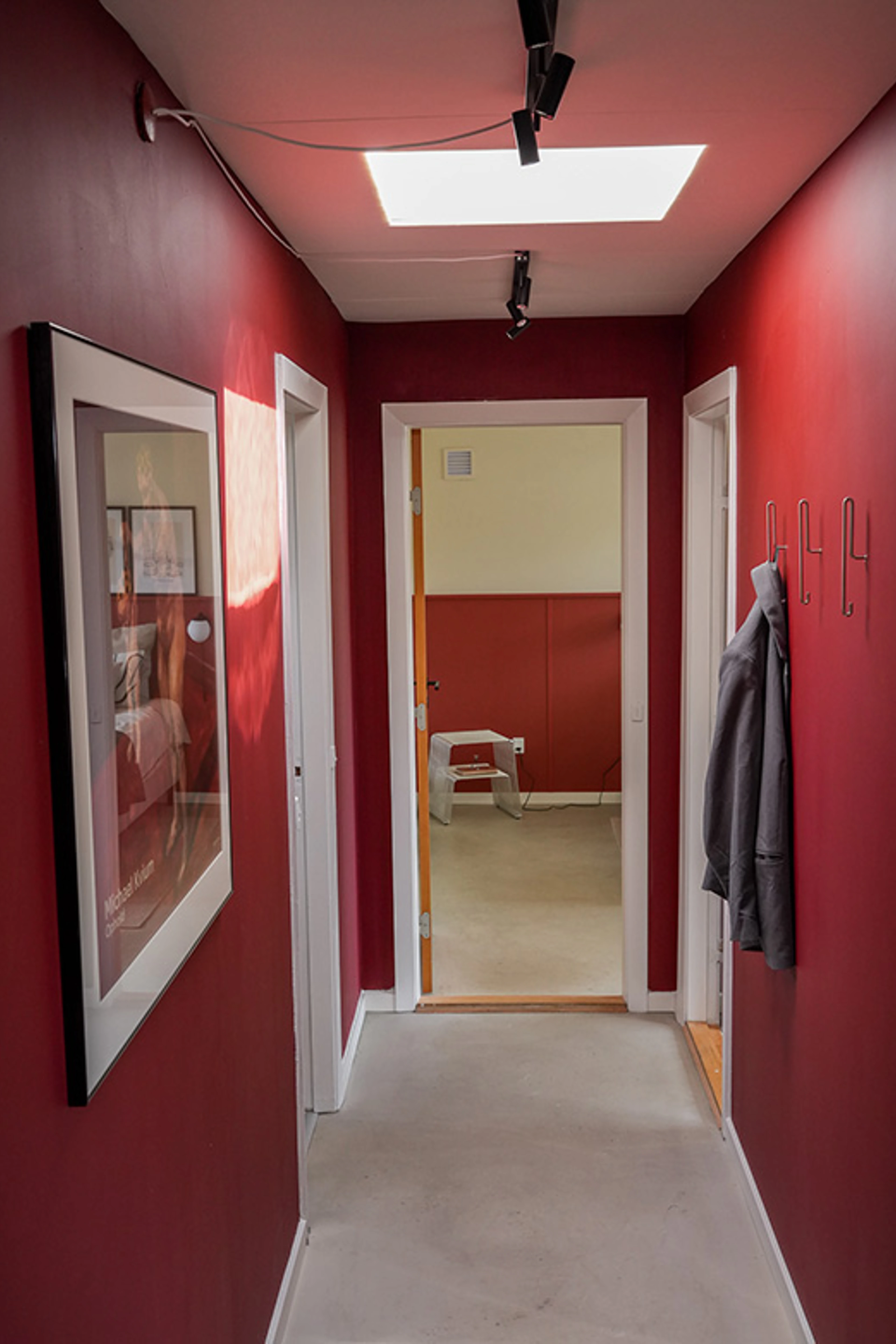 Smal gang med vægge malet i en dyb rød nuance. En indrammet plakat hænger på den ene væg, mens knager med ophængt overtøj og spotbelysning fremhæver rummets farverige udtryk.