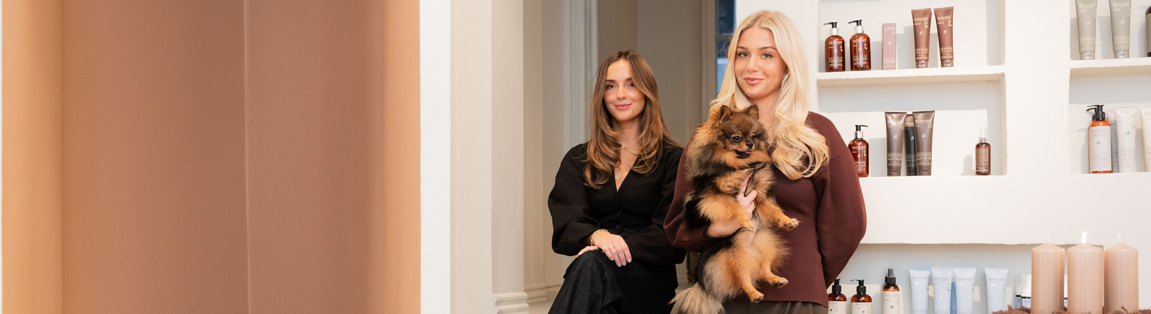 To kvinder i frisørsalon, hvor den ene holder en lille hund foran hylder med hårprodukter.