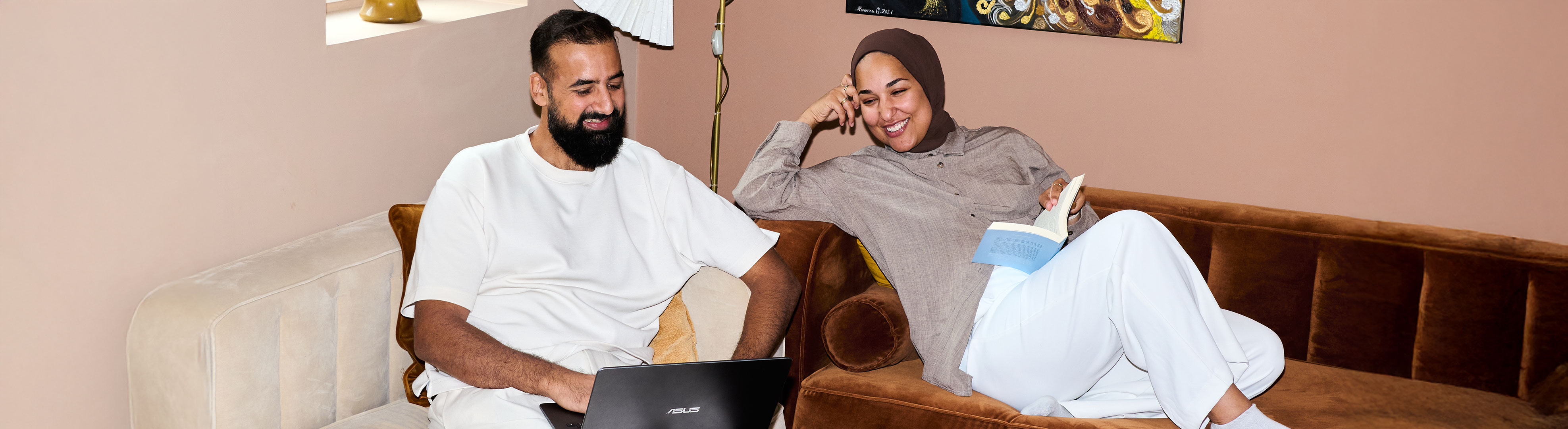 Two people sit relaxed on a sofa, one works on a laptop while the other smiles with an open book in hand.