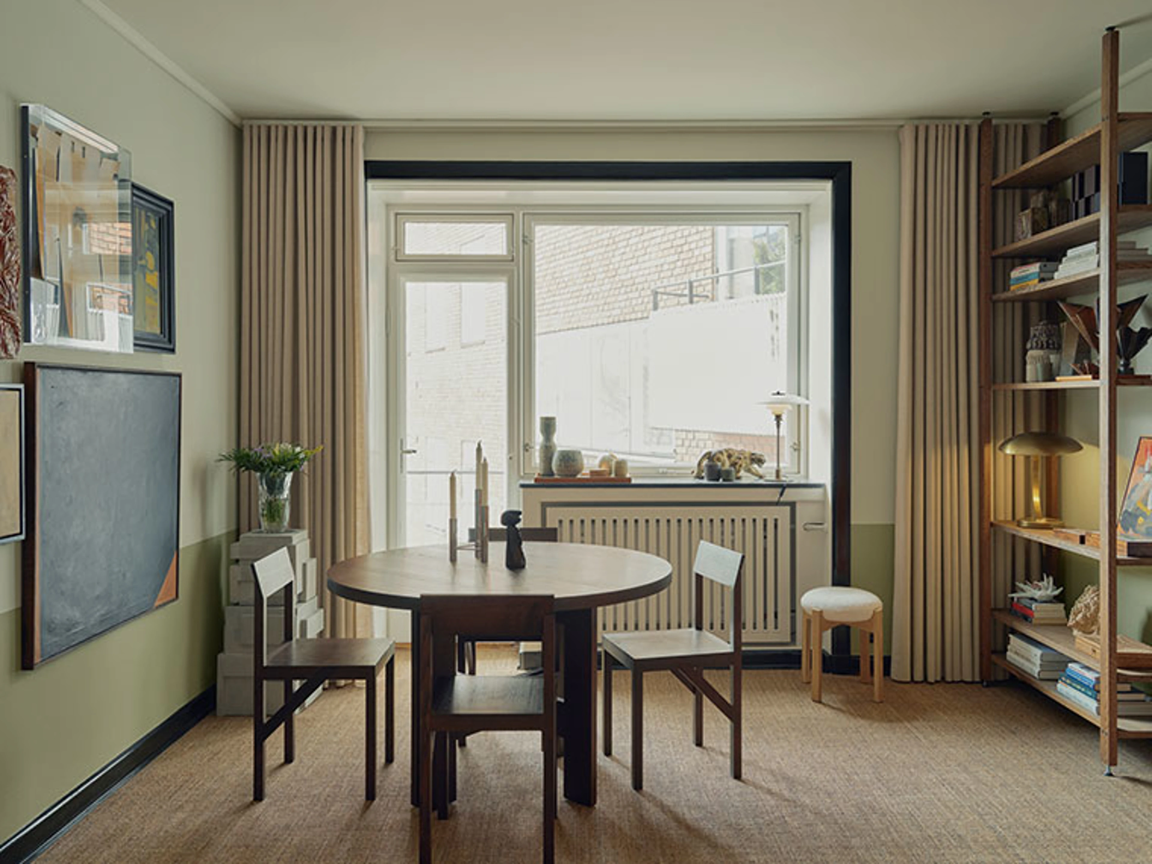 Dining room with a round dark wood table, simple chairs, large windows, long curtains, and artwork on the walls.
