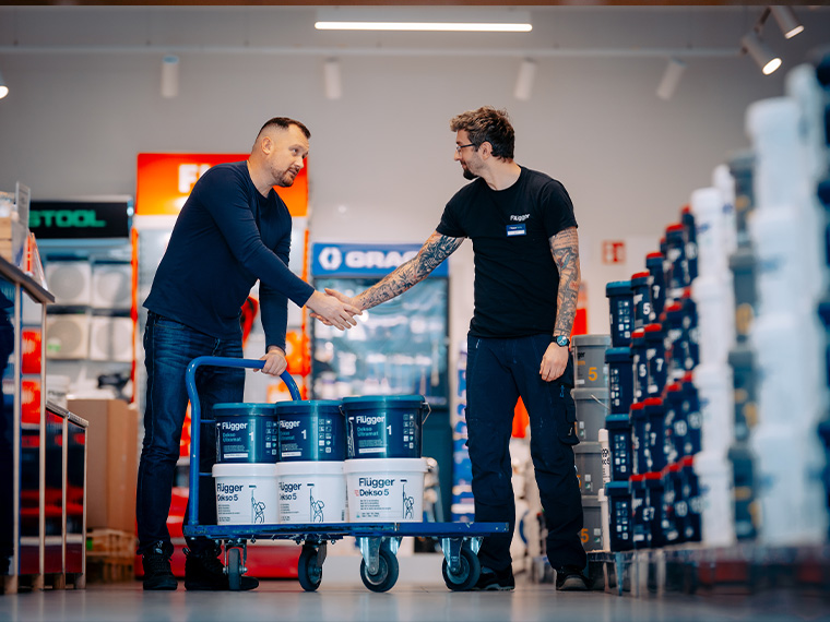 Two people shake hands in a Flügger store next to a cart with paint buckets. Shelves of paint and painting supplies are visible in the background.