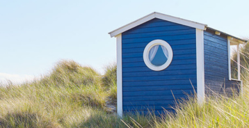 Blå trästuga vid stranden med ett runt fönster, omgiven av dyngräs.