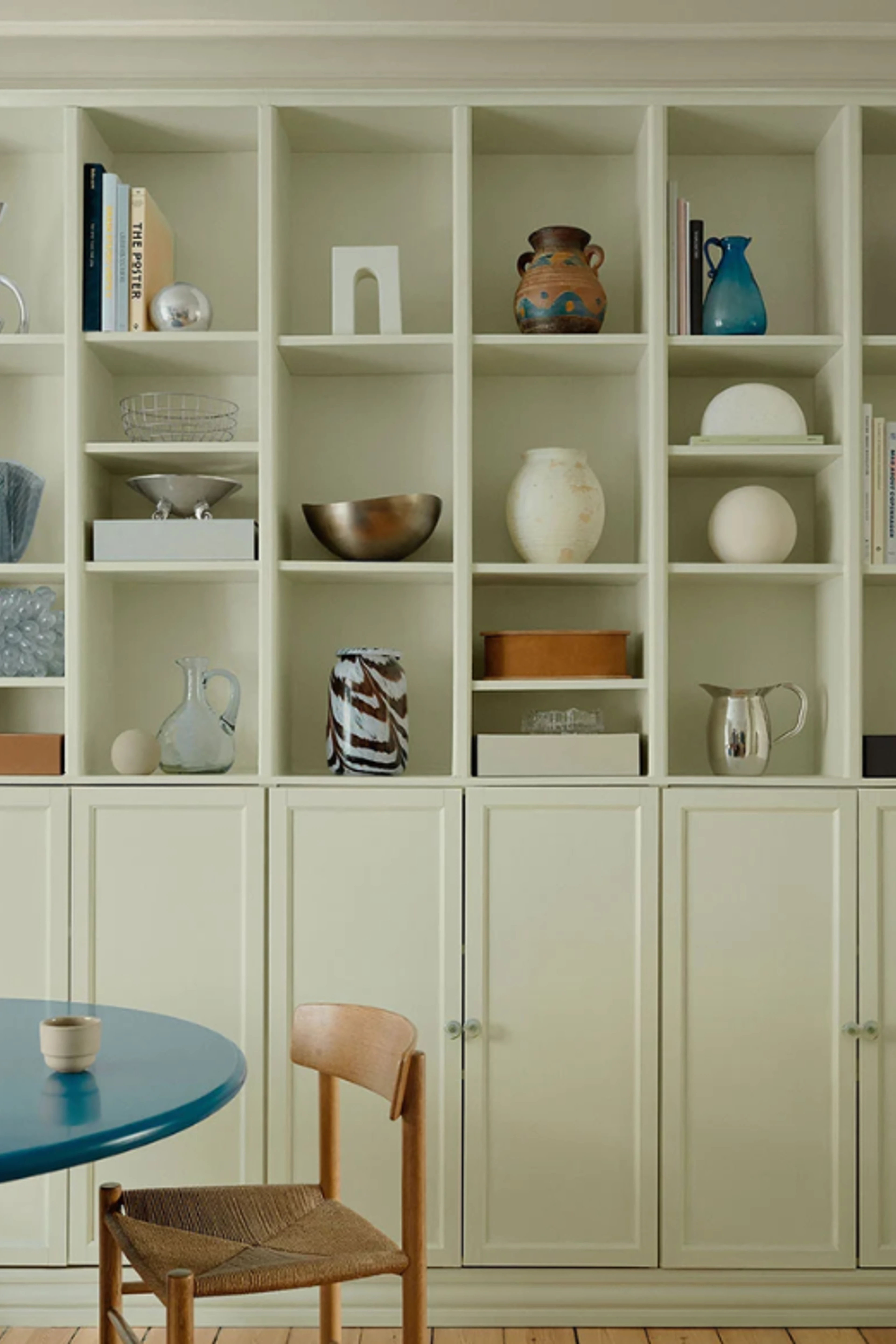 Beige reol med åbne hylder fyldt med keramik, glas, skåle og bøger i rolige, neutrale farver.