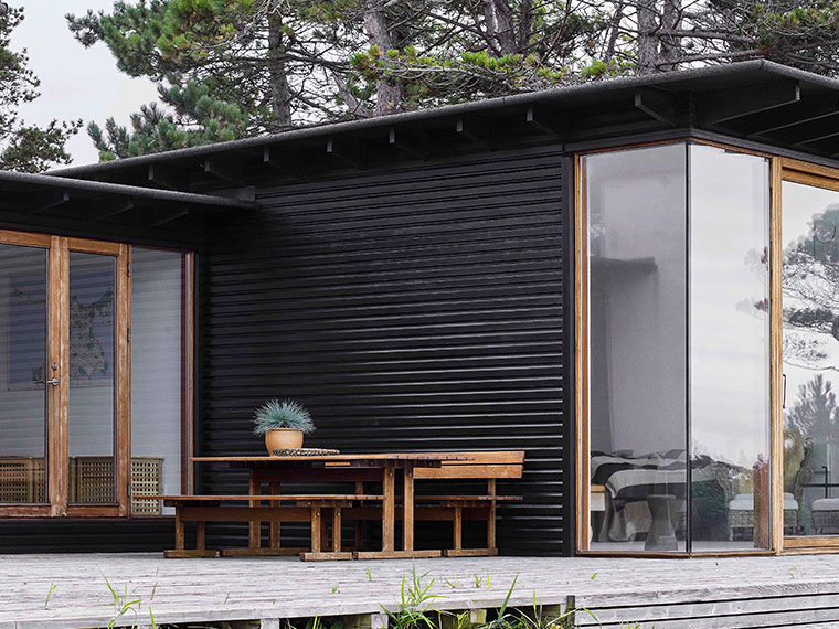 Svartmalt trehus med kledning, treterrasse med spisebord og benker, omgitt av bartrær.