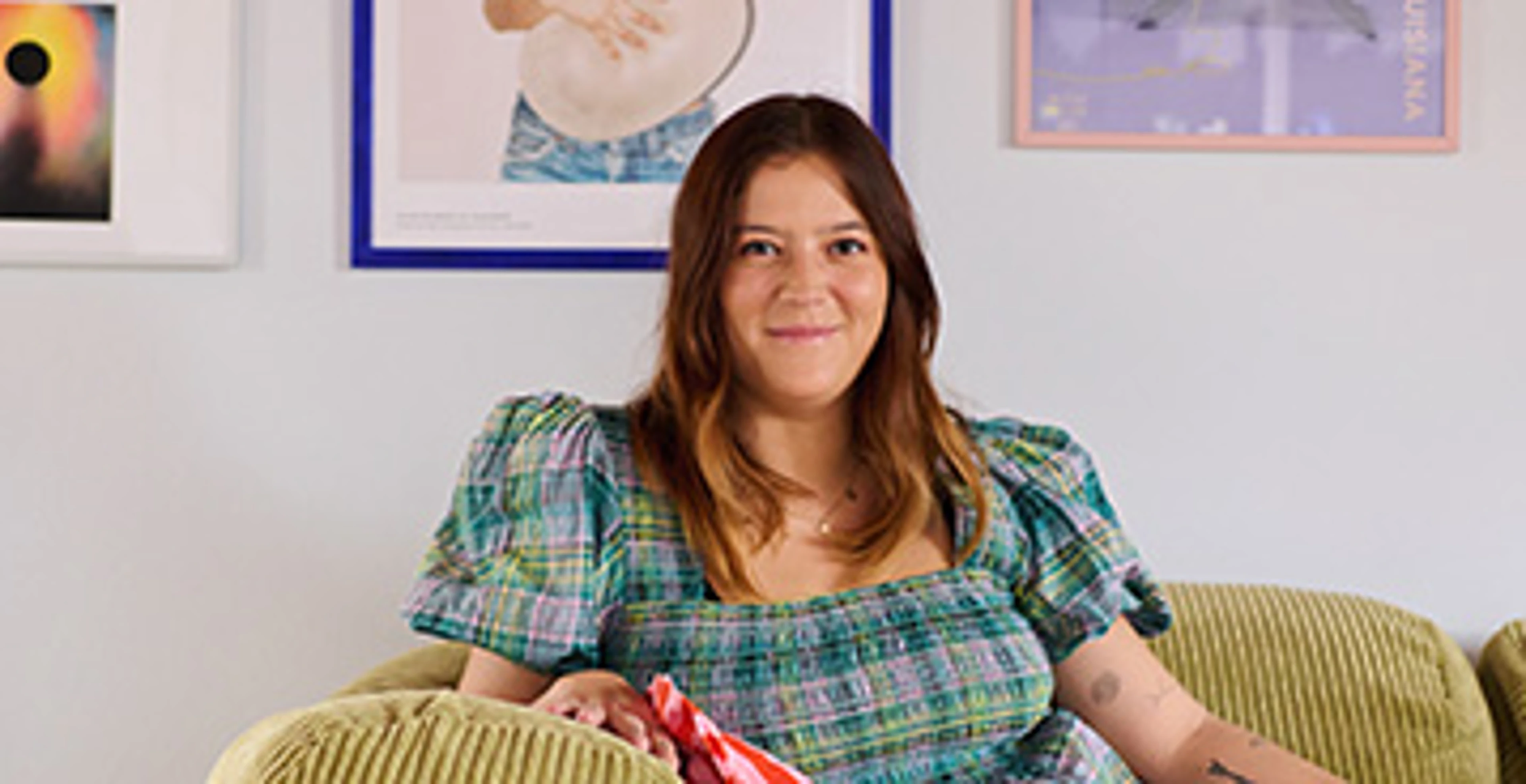 Belinda kigger ind i kameraet mens hun læner sig op armlænet på sofaen.