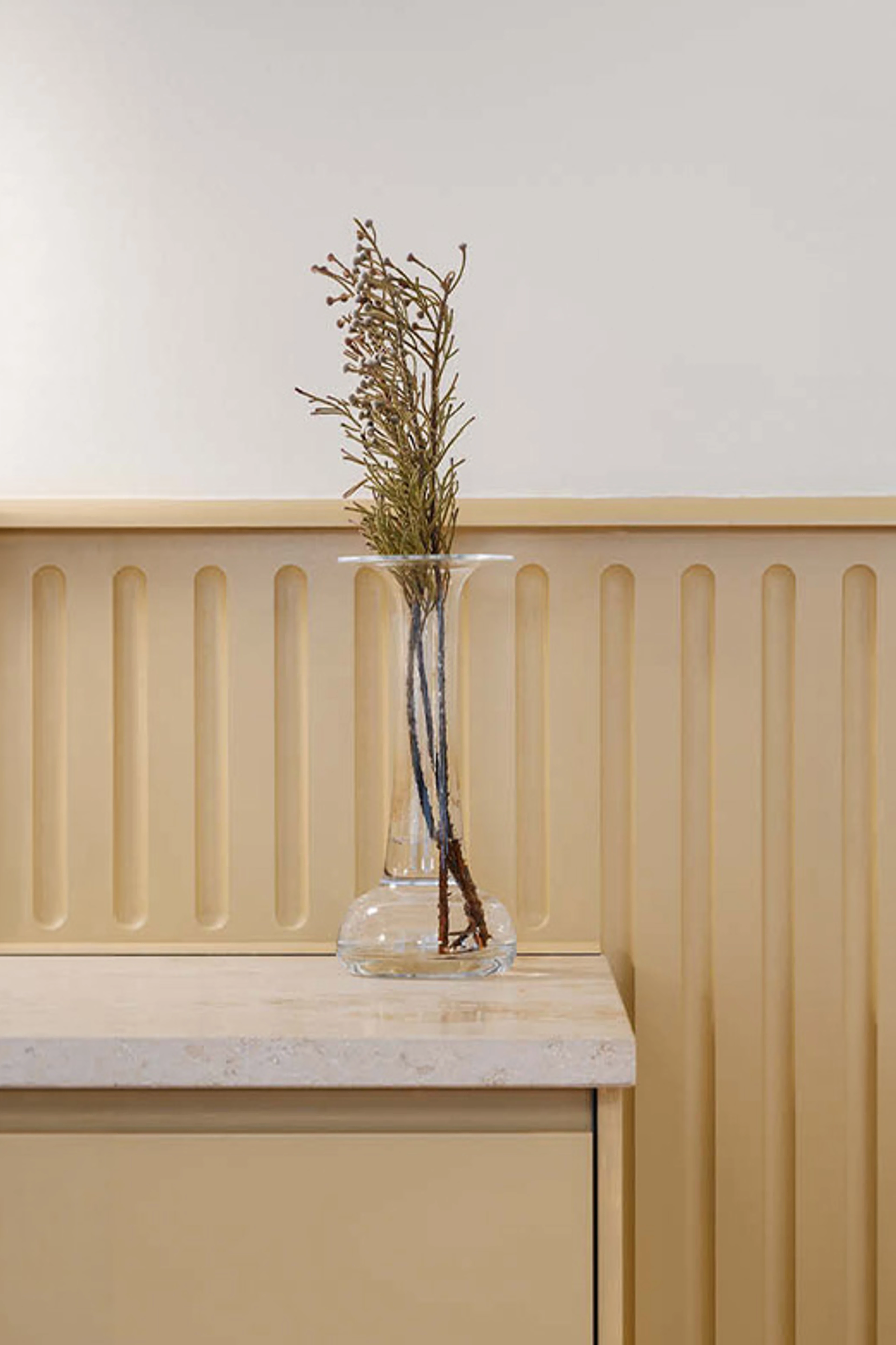 Beige panelvegg med glassvase og tørkede grener på et lyst skap.