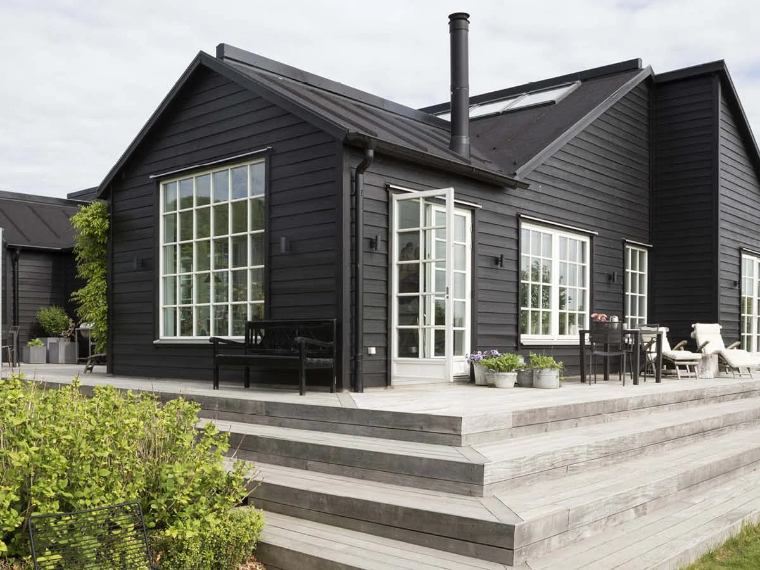 Svart trähus med vita fönsterkarmar, ett stort trädäck med trappsteg och trädgårdsmöbler.