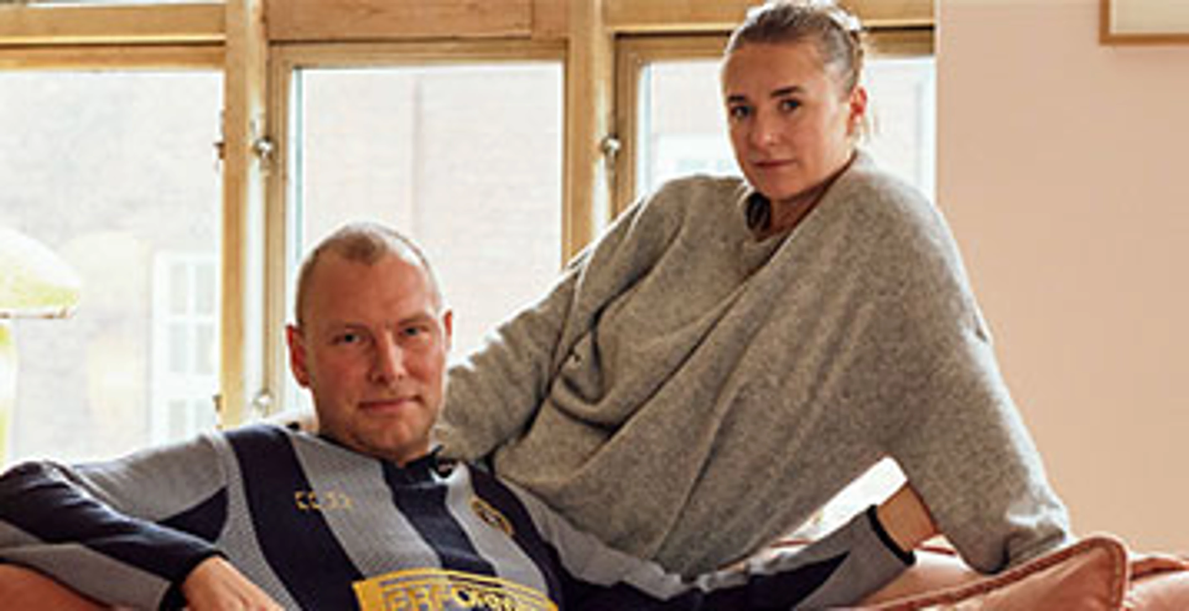 Par sidder tæt sammen på lyserød sofa foran store vinduer.