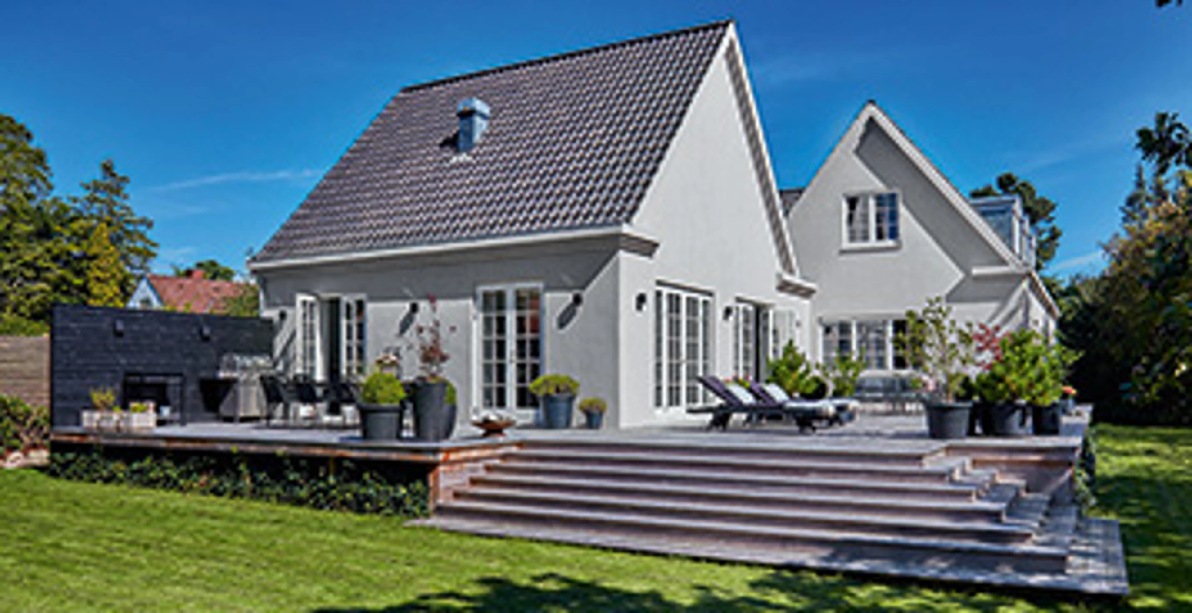 Hus med gråmalt fasade. Det er en treterrasse rundt huset, og en grønn gressplen i forgrunnen.