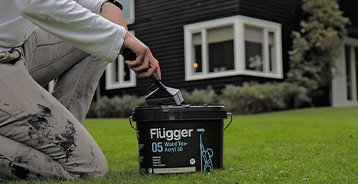 Person knæler på en græsplæne med en spand Flügger Wood Tex 05 Acryl 30 træbeskyttelse og en pensel. I baggrunden ses et sortmalet træhus med hvide vinduesrammer.