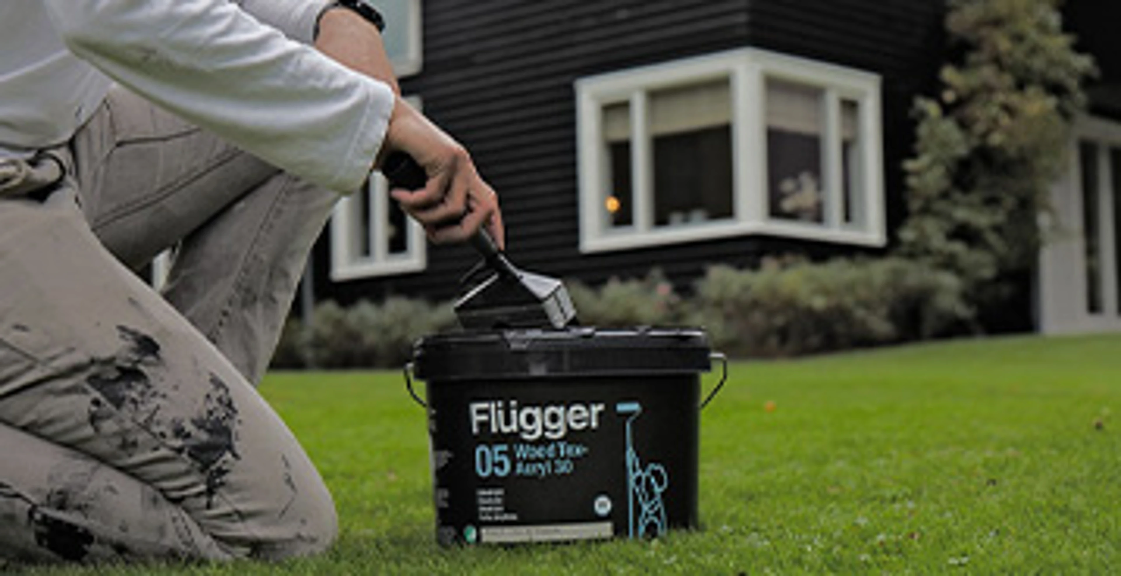 Person knæler på en græsplæne med en spand Flügger Wood Tex 05 Acryl 30 træbeskyttelse og en pensel. I baggrunden ses et sortmalet træhus med hvide vinduesrammer.